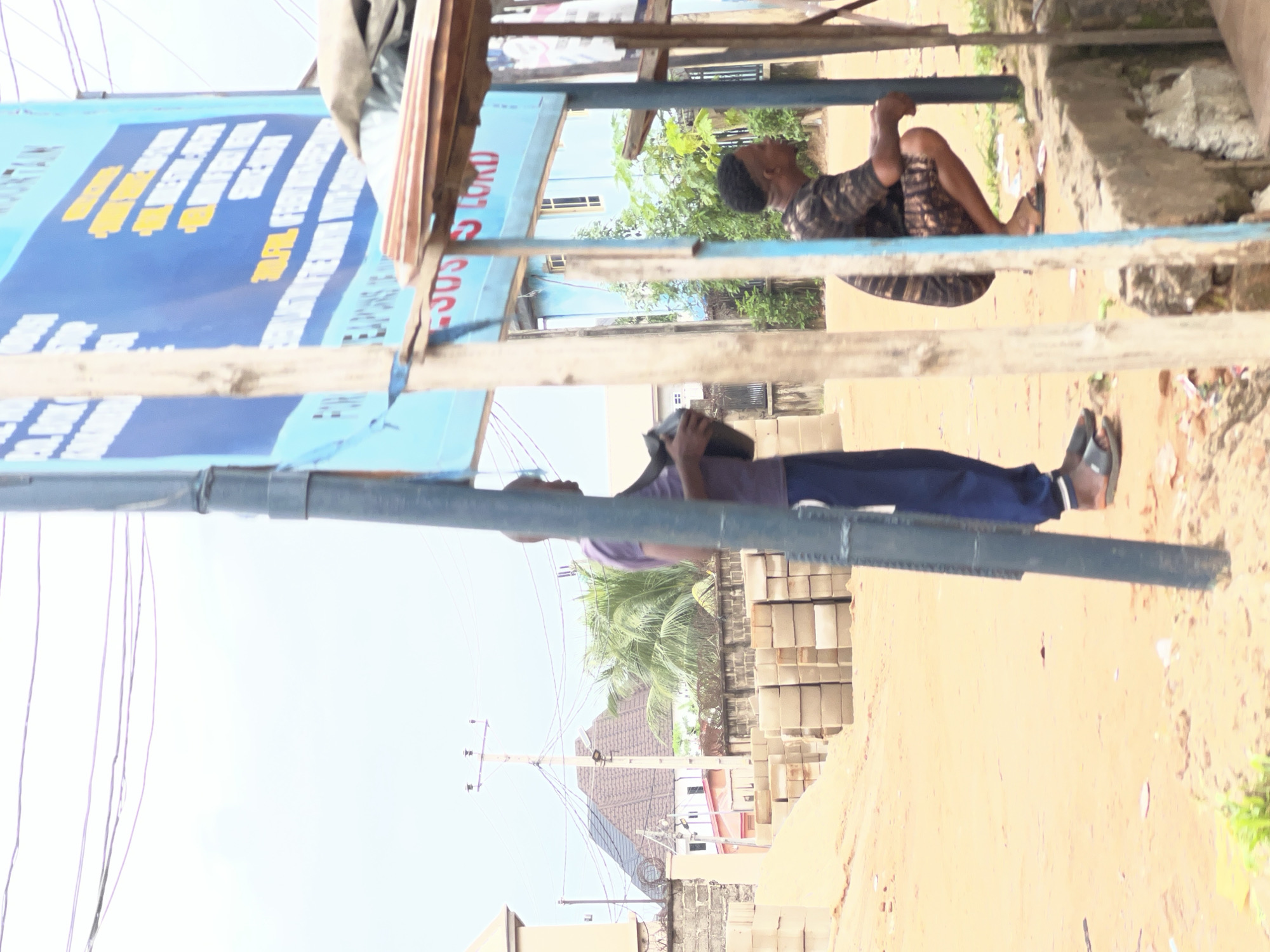 In Nkpor, Nigeria, on a dusty, unpaved street, a scene unfolds near a roadside stall. A large blue sign with white and yellow text is mounted on a wooden post, suggesting a religious or community announcement. Below the sign, a makeshift stall constructed from wood and corrugated metal is visible, with a person in a dark, patterned short-sleeved shirt and shorts squatting nearby. Another person, wearing a purple shirt and dark blue trousers, stands holding a dark bag. In the background, stacks of grey concrete blocks lie on the ground, indicating construction or development. Beyond the blocks, buildings with pitched roofs and palm trees are visible, along with electrical poles and wires crisscrossing the sky. The ground is primarily sandy and littered with debris.