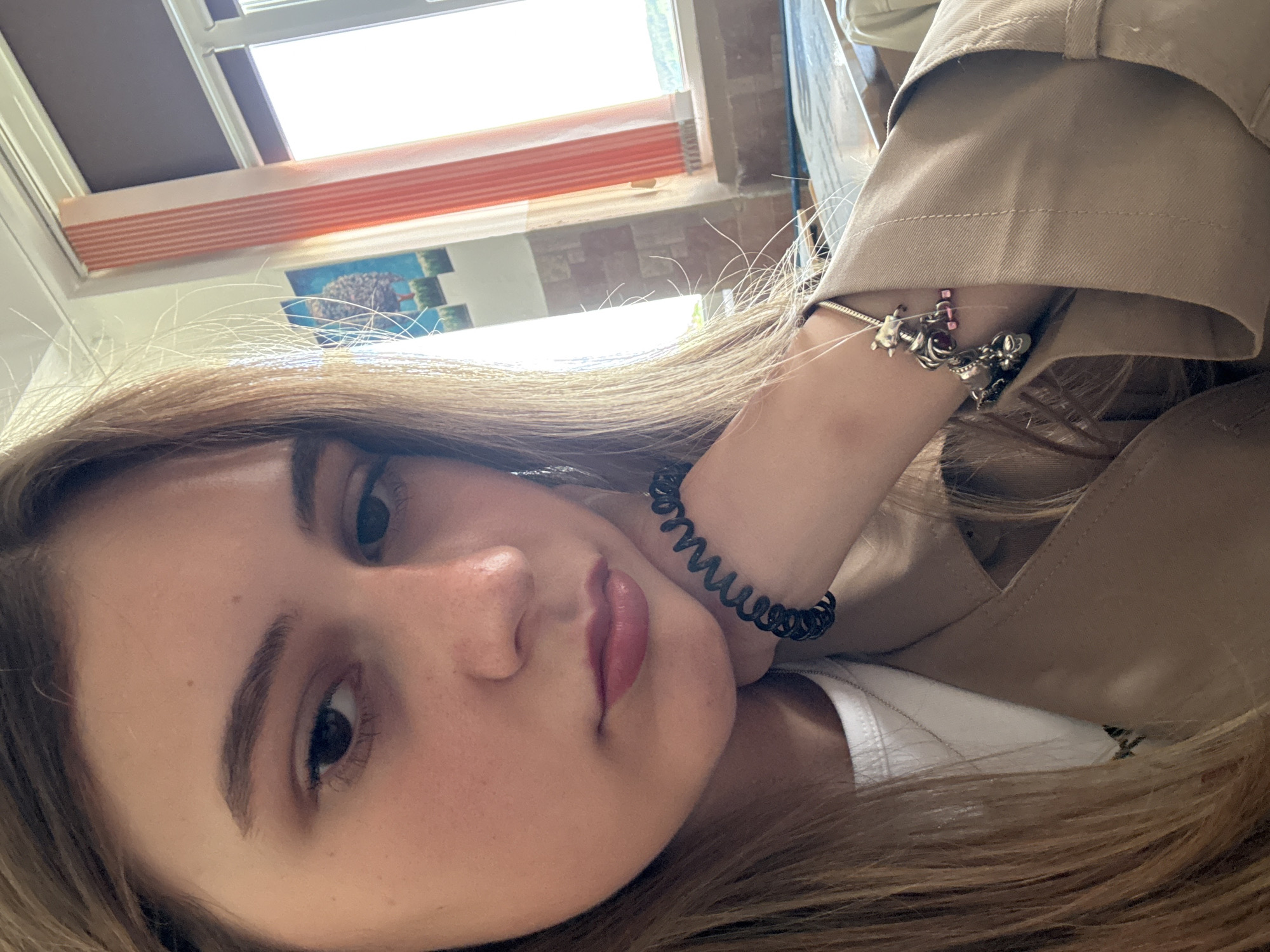 A close-up, selfie-style image shows a young woman with light brown, shoulder-length hair. She is fair-skinned with brown eyes and light eyebrows. She is wearing a light tan trench coat over a white top. On her right wrist are two bracelets: a coiled black hair tie and a charm bracelet. The background includes a window with orange and white blinds and a glimpse of a picture on the wall. Without external data, it is impossible to identify if the location is in Panagyurishte, Bulgaria.