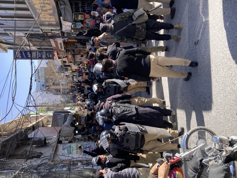 The image displays a densely populated street scene in Quetta City Tehsil, Pakistan, under clear daylight with strong shadows. The street is flanked by multi-story commercial buildings featuring numerous shopfronts with signage in Urdu and English, including "IQBAL STATIONERY MART" and "EPCOT." A complex network of overhead electrical and communication wires spans the street. Mountains are visible in the background under a clear blue sky.
In the foreground and midground, a contingent of approximately fifteen to twenty law enforcement personnel is present. Several individuals are equipped with black tactical vests, helmets (some with visors), khaki trousers, and carry batons and clear riot shields marked "POLICE." Other officers wear khaki trousers and dark long-sleeved shirts. One individual on the right wears camouflage trousers, a dark top, and carries a long-barreled firearm, possibly a shotgun, slung over their shoulder, along with a canvas bag. Most law enforcement personnel face away from the camera, forming a line or cordon.
Behind the law enforcement personnel, the street extends into a crowded commercial area filled with a large number of civilians, predominantly males, dressed in a mix of traditional Pakistani attire and Western casual wear. They appear to be moving through or congregating within the market. A red and chrome motorcycle is partially visible in the bottom left corner of the frame.