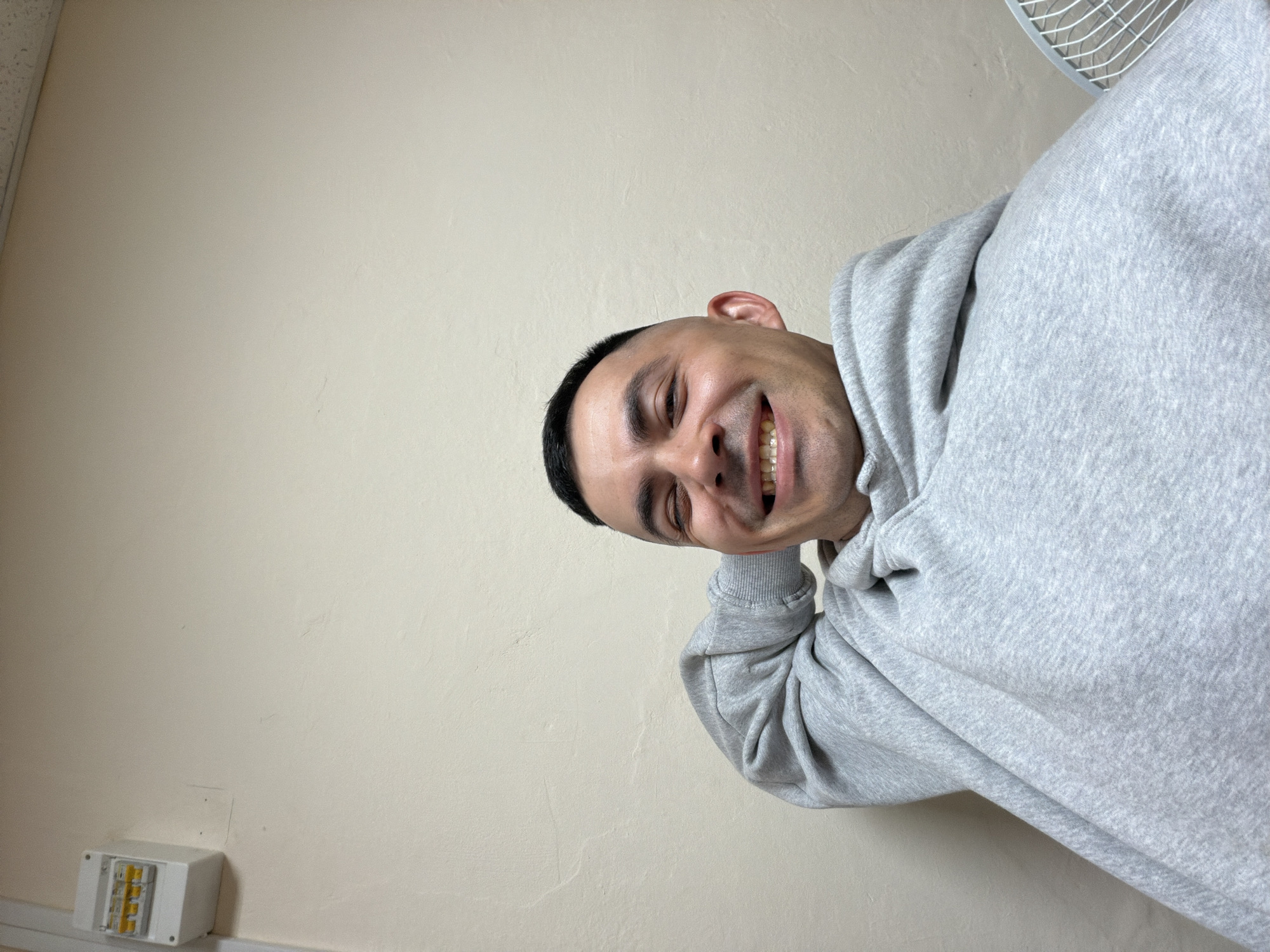A person with dark hair and a gray hoodie is smiling at the camera. The background is a plain, light-colored wall with an electrical box in the lower-left corner. A fan is visible in the upper-right corner. The location is not identifiable as Izhevsk, Russia.
