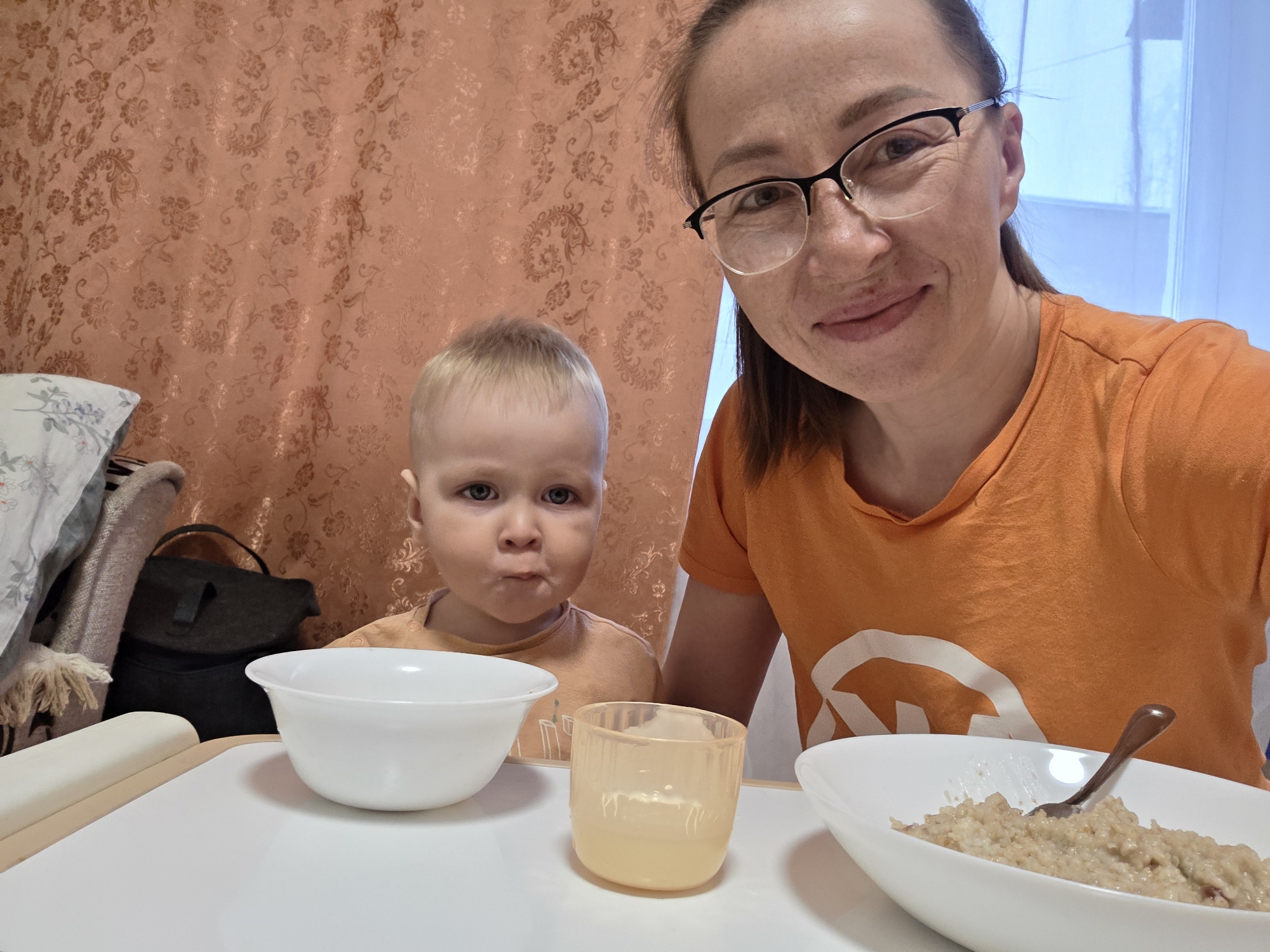 This indoor photograph captures a candid selfie moment between a woman and a young child during what appears to be a mealtime.
In the foreground, a white table or high chair tray is visible, holding several dishes. To the left, there's an empty white bowl and a translucent yellowish-orange cup with some liquid, positioned in front of the child. To the right, in front of the woman, is a white elongated bowl filled with a light brown, thick substance resembling oatmeal or porridge, with a dark spoon resting in it.
The subjects are a woman, who is taking the selfie, and a young child. The woman, positioned on the right side of the frame, has dark hair pulled back, wears glasses, and is smiling subtly. She is wearing a bright orange t-shirt with a partial white graphic visible on the chest. The child, a toddler with blonde hair and blue eyes, is on the left side of the frame. The child is making a distinctive expression, with puffed cheeks or a slight pout, and is also wearing an orange t-shirt.
The background features a wall or curtain with a peach or light orange color and an intricate, textured floral pattern. To the far right, a bright light blue area suggests a window or an adjacent room. On the left side of the image, behind the child, parts of a white pillow with a blue floral design, a textured beige blanket, and a dark bag are visible.
The overall setting appears to be a domestic one, likely a kitchen or dining area. The lighting suggests it is daytime. There is no clearly decipherable text visible in the image, apart from the partial graphic on the woman's shirt.