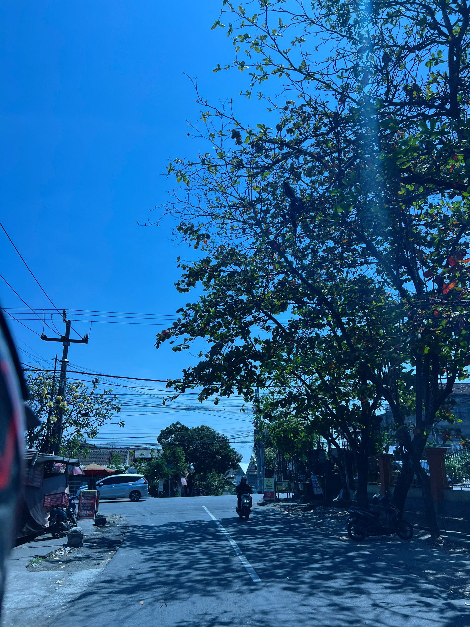 The image captures a street scene in Denpasar, Indonesia, under a clear blue sky. In the foreground, the left side of the frame is occupied by the blurred edge of a vehicle, likely a car, reflecting the bright sunlight. A paved road stretches into the distance, with a white line marking the center.
On the left side of the road, there's a small roadside stall with a red awning and a motorbike parked nearby. A silver van is visible further down the street, parked on the side. A power pole with numerous wires crisscrosses the sky on the left.
A single individual on a motorbike is heading away from the viewer down the road. On the right side of the road, a large tree with lush green leaves casts dappled shadows onto the asphalt. Behind the tree, there are glimpses of buildings and a fence.
The overall scene suggests a typical day in a bustling Indonesian city, with evident infrastructure of power lines and the presence of motorbikes, a common mode of transport. The bright sunlight indicates it's daytime, and the clear sky suggests fair weather.