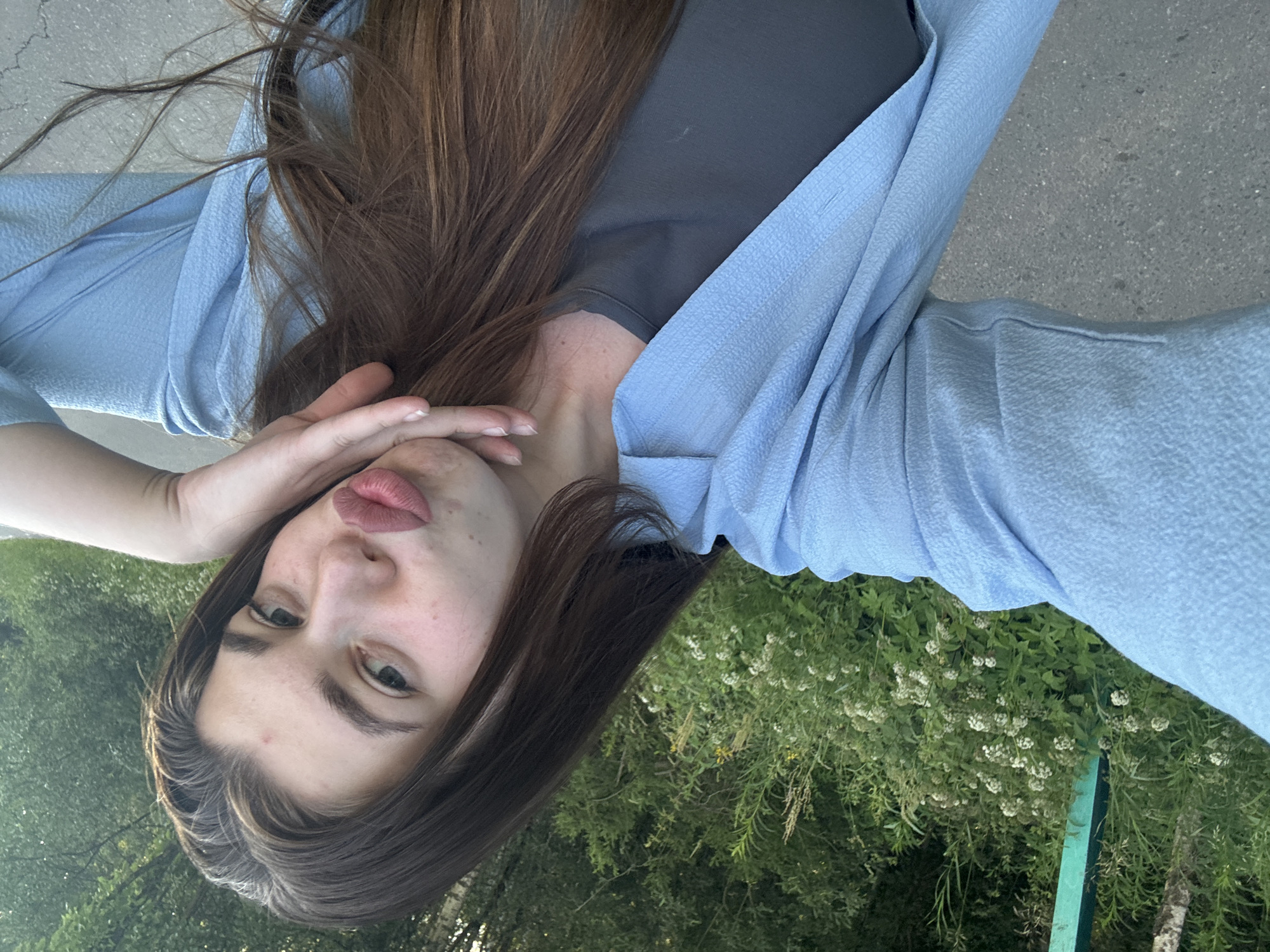 This is a selfie of a young woman in an outdoor setting, likely in Шевлягино, Russia. The image is oriented sideways, with the woman's head towards the bottom of the frame. She has long, brown hair and is wearing a dark grey t-shirt and a light blue, textured open cardigan or jacket. Her left hand is raised to her face, partially covering her mouth, and her lips are slightly parted, giving her a pouting or playful expression. Her eyes are looking towards the camera with a neutral to slightly curious gaze.
The background consists of lush green foliage, suggesting a natural environment with trees and plants. There are some small white flowers visible among the greenery, and a portion of a light grey paved surface, possibly a sidewalk or path, is also in view. A thin, bright green vertical object, possibly a post or railing, is visible on the right side of the frame. The lighting suggests it is daytime, and the outdoor setting with green vegetation indicates pleasant weather. The overall mood of the image is casual and relaxed. There is no visible text in the media file.