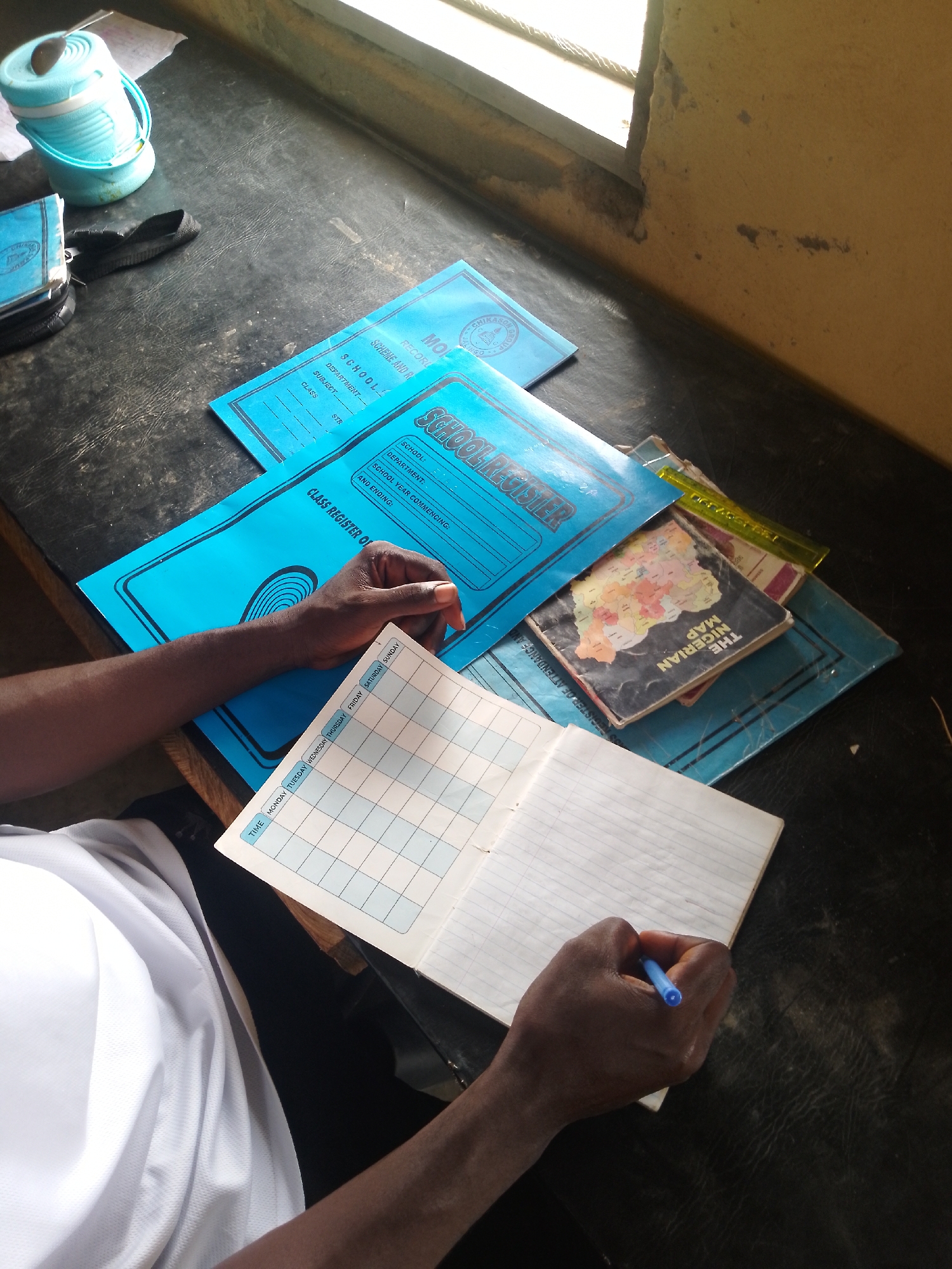 In an indoor scene, a person's dark-skinned arm and hand are visible, holding a blue pen and writing in an open notebook. The notebook lies on a dark, textured surface, likely a desk. Other objects on the desk include several blue school registers, one of which is partially open and displays text such as "SCHOOL REGISTER." There is also a closed book with "THE NIGERIAN MAP" visible on its cover, and a yellow ruler. To the left, a light blue thermos with a dark lid and handle sits on a white piece of paper. The background shows a window with natural light entering and a section of a yellow-painted wall with some damage. The environment suggests a classroom or study area.