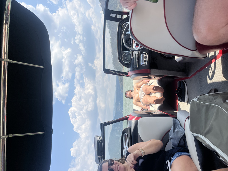 The image presents an outdoor scene aboard a recreational boat on a large body of water, identified as being in or near the city of El Cerrillo, Mexico. The environment consists of calm, greenish-blue water under a clear blue sky with scattered white cumulus clouds, bordered by distant, tree-covered hills on the horizon.
Three individuals are present. In the left foreground, a woman with dark hair, sunglasses, and a black tank top is seated, smiling. Her left arm displays multiple bracelets, and a white object, possibly a phone, rests on her lap near blue denim shorts and a grey fabric item. Centrally positioned in the bow of the boat, a young male lies supine and shirtless on a padded, white and red sun deck, wearing light-colored swim trunks, with his bare feet visible, apparently sunbathing. To the right foreground, the arm and leg of a third person are partially visible, seated in a white and red upholstered boat chair, holding a green bottle.
The boat's interior features include white and red upholstered seats, a black dashboard with gauges and a steering wheel on the starboard side, and a windshield. A black fabric bimini top provides overhead cover. On the boat's grey floor, a pair of black athletic shoes are situated between the sunbathing male and the helm, while a green and grey duffel bag rests in the left foreground near the seated woman.