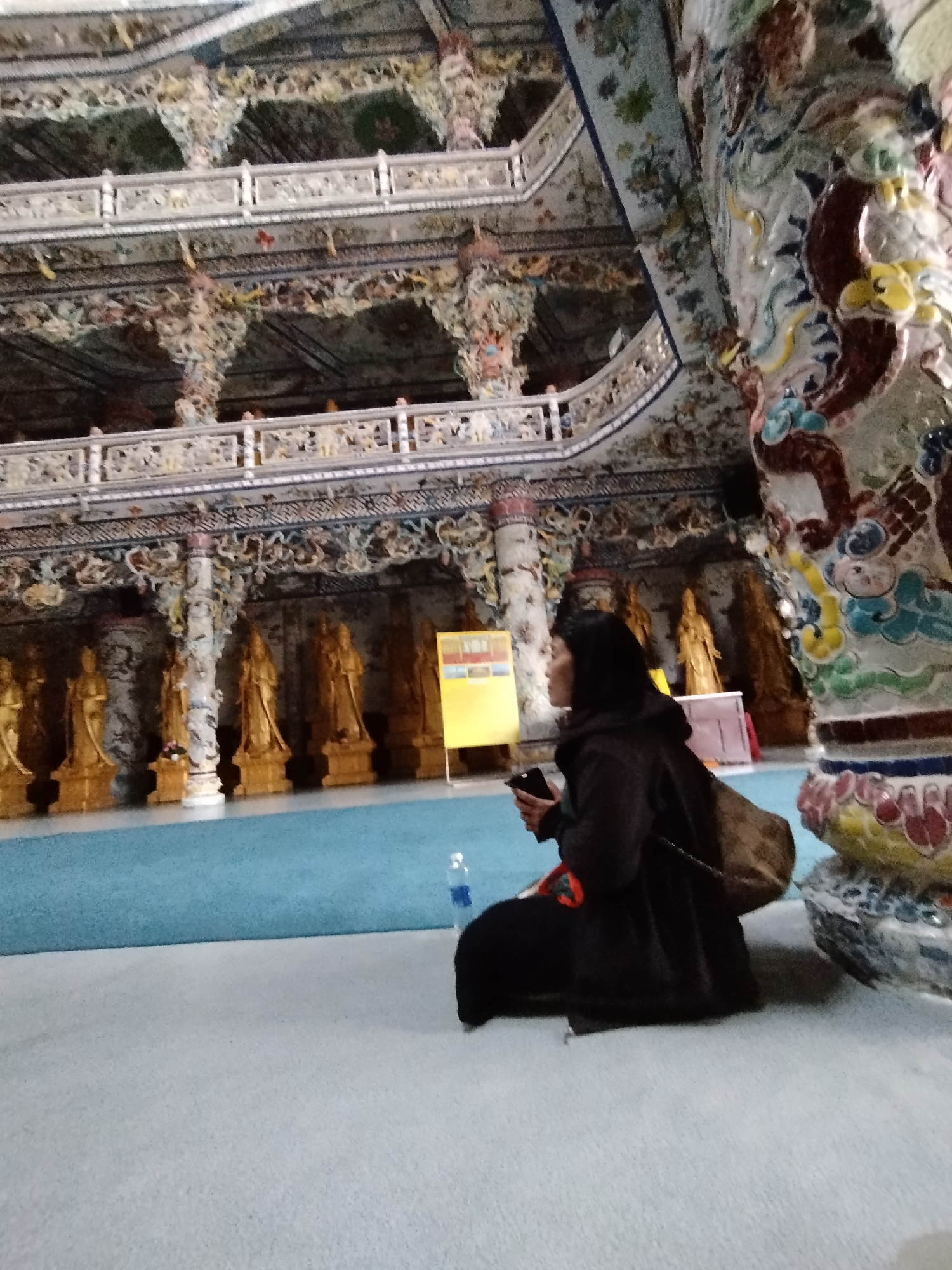 A woman in a black hooded coat sits cross-legged on a light-grey floor in a large, ornately decorated building, possibly a temple, in Xuân Thành, Vietnam. She holds a dark object in her hand, possibly a phone. A clear plastic water bottle sits on the blue floor in front of her. Behind her are rows of gold-colored statues, and a yellow sign. Elaborate carvings and architectural details adorn the walls, ceilings, and columns of the building.