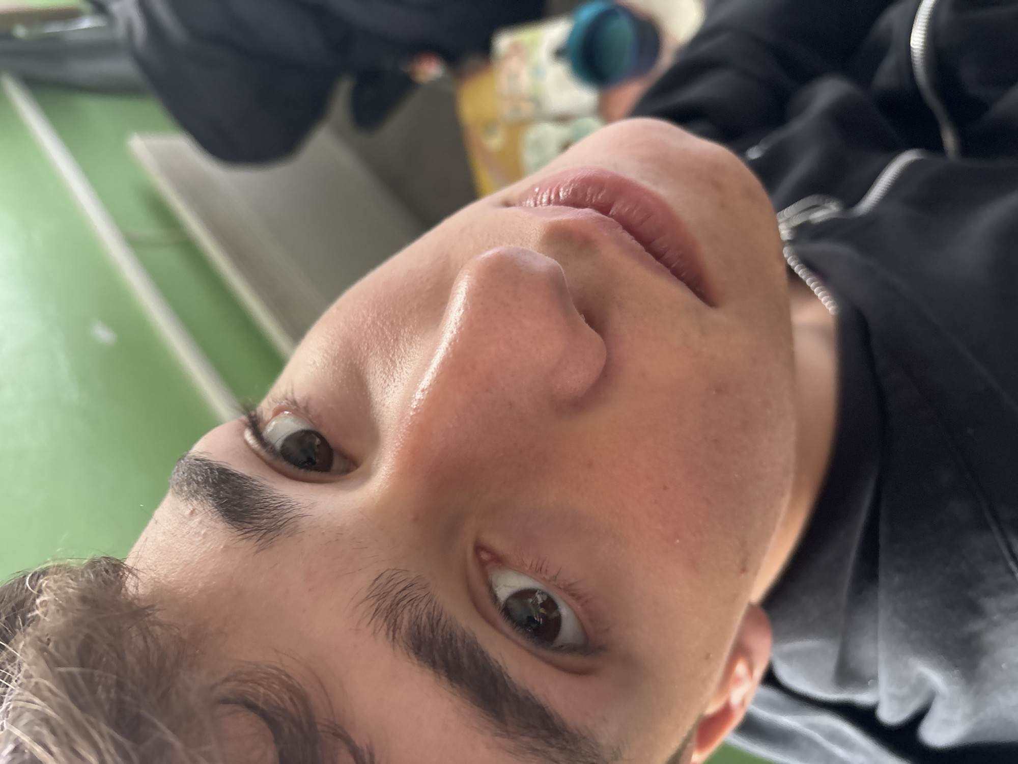 The image displays a close-up, upward-angled portrait of a young male subject situated in an indoor setting within Troyan, Bulgaria. The individual occupies the majority of the frame, presenting light skin, dark brown eyes, and short, wavy brown hair on the forehead. His dark eyebrows are distinct, and his lips are slightly parted. Minor skin textures and pores are visible due to the proximity. He is dressed in a dark, likely black, zippered garment, with a metallic zipper pull partially discernible at the bottom right. The background on the upper left features a vibrant green wall with a prominent horizontal light-colored stripe or trim. Further to the right in the blurred background, a dark, indistinct shape, a yellow object (possibly a book or box), and a blue cup containing liquid are visible. The subject's gaze is directed slightly above and to the right of the camera's apparent axis.
