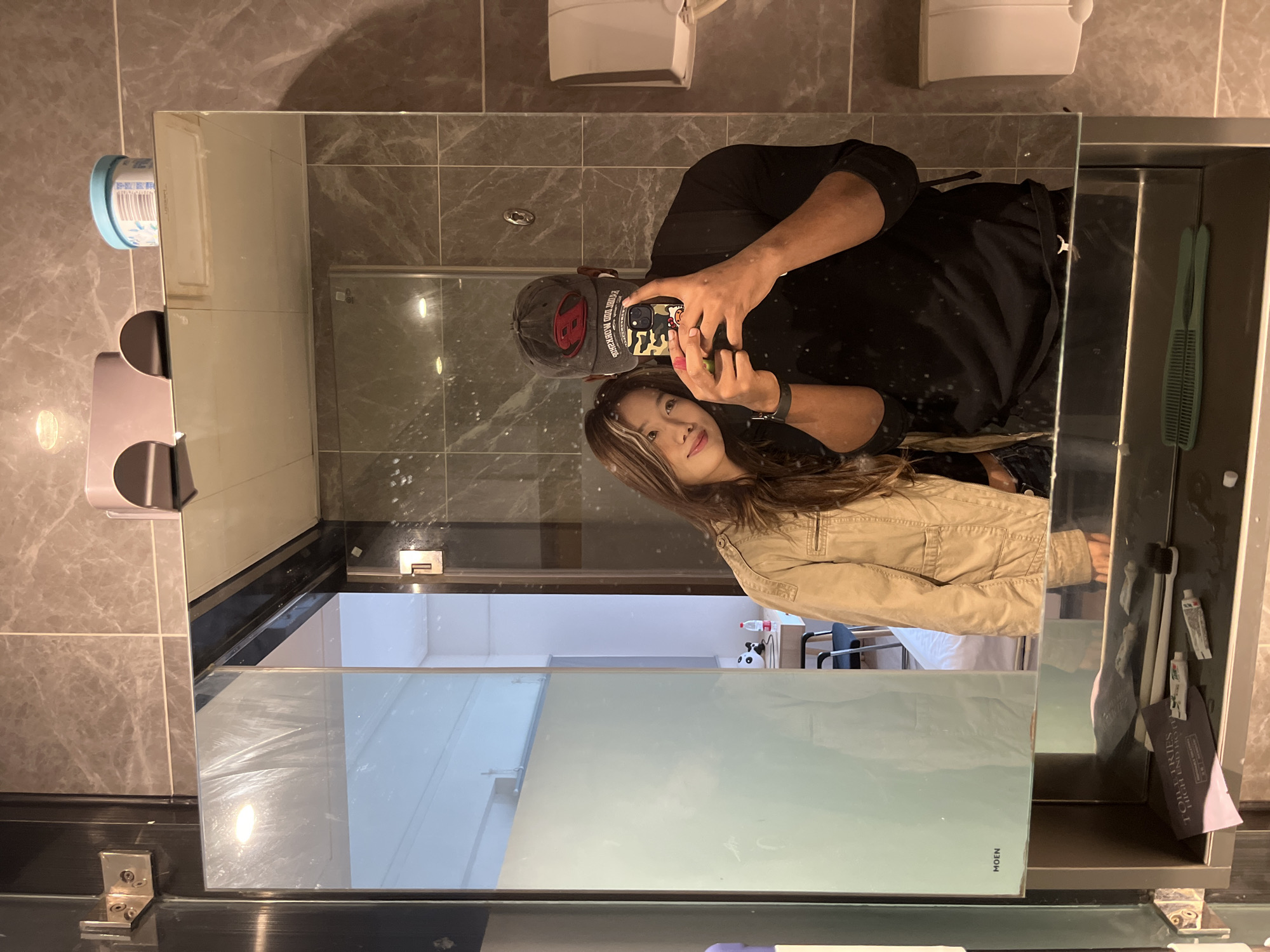The image shows a selfie taken in a bathroom setting. A woman with long brown hair is standing next to a person wearing a black t-shirt and a baseball cap, who is holding a smartphone to take the picture in the mirror. The mirror reflects the bathroom, including gray tiled walls, a toilet paper dispenser, and a vanity area with toiletries. The reflection shows a glimpse of a white room, with chairs. Without additional visual or contextual evidence, the precise location within the Chenghua District of China cannot be determined.