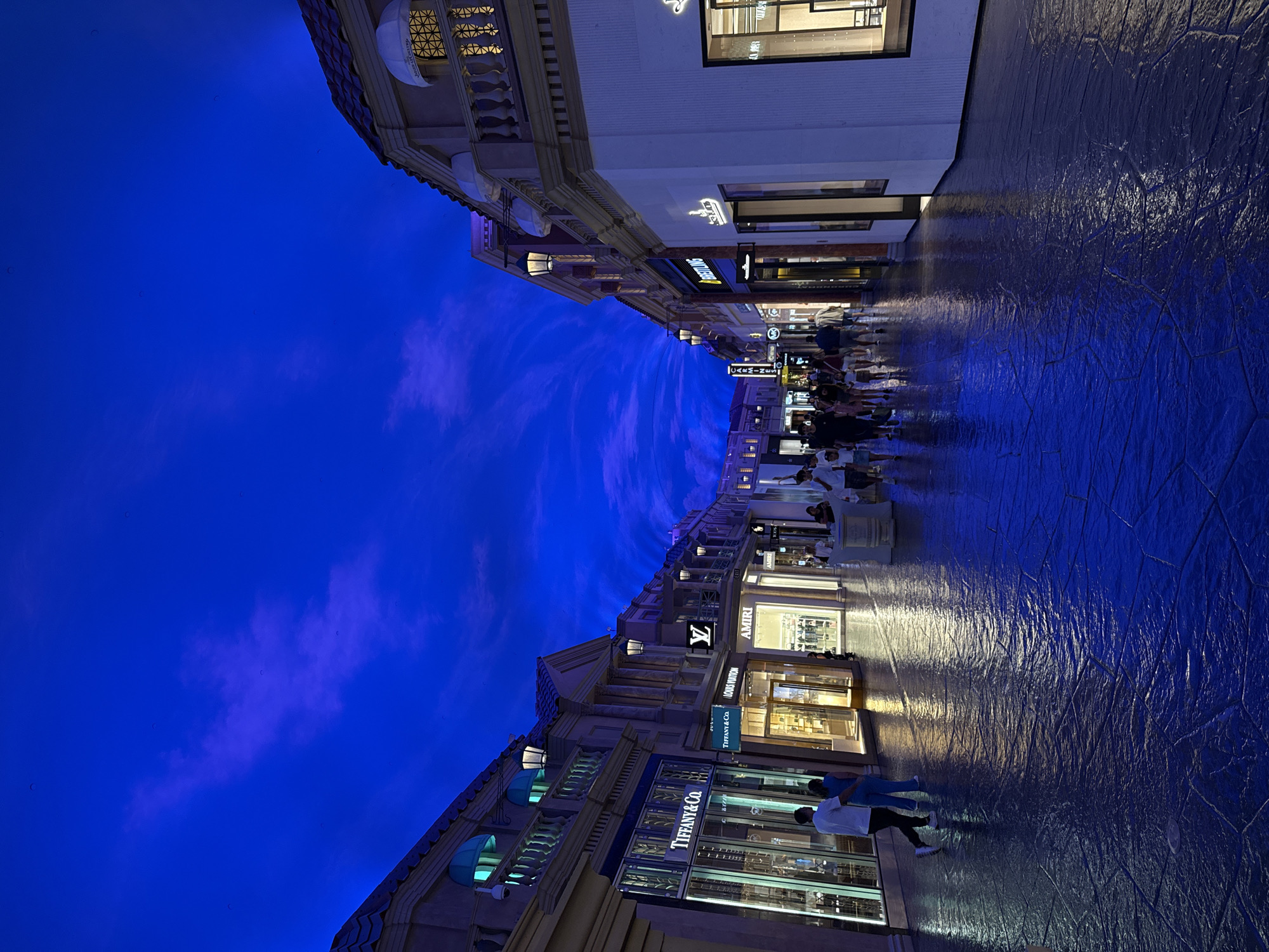 The image depicts an indoor shopping area with a ceiling designed to mimic a twilight sky with clouds. The architecture features classical elements, reminiscent of European streetscapes, with storefronts lining both sides of a wide walkway. The floor is paved with large, irregular stone tiles that are wet, reflecting the vibrant blue of the ceiling and the lights from the shops.
Several people are visible walking along the path. On the left, two individuals are walking away from the camera, one wearing a white t-shirt and the other in blue attire. Further down the walkway, a group of people are seen browsing or walking. The storefronts include well-known luxury brands such as Tiffany & Co., Louis Vuitton, and Amiri, indicated by illuminated signage. Other shops with less distinct signage are also present. Streetlights are mounted along the buildings, casting a warm glow that contrasts with the cool blue of the ceiling. The overall impression is a simulated outdoor environment within a controlled indoor space, likely a high-end shopping mall or resort.
This scene is located at The Forum Shops at Caesars Palace in Paradise, Nevada, United States of America.