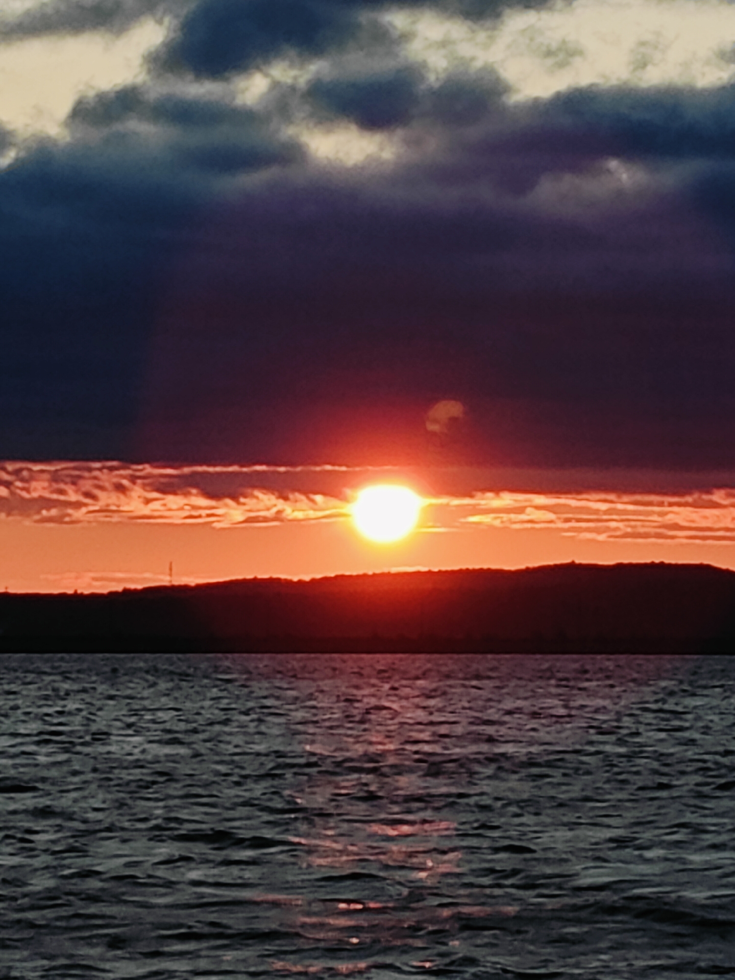 The image depicts a sunset over a body of water, likely the Kama River in Chaikovsky, Russia, given the silhouette of hills that are characteristic of the area. The sun is a bright, glowing orb near the horizon, casting a warm orange and red light that illuminates the lower portion of the sky and reflects on the rippling water. Dark, heavy clouds dominate the upper sky, creating a dramatic contrast with the vibrant sunset. The water is dark and choppy, with streaks of reddish light from the sunset visible on its surface. The landscape is reduced to a dark silhouette against the colorful sky. There are no people or man-made structures clearly visible, except for a faint, thin vertical structure on the horizon line. The overall scene is serene and natural, with the dramatic sky adding a sense of awe.