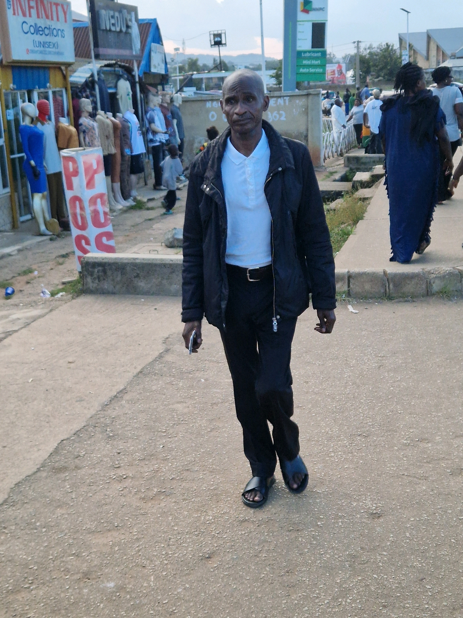 An outdoor medium shot captures a bald, dark-skinned man walking towards the camera on a paved street in a commercial area. He is wearing a white collared shirt, a black jacket, black pants, and black sandals. His left hand is extended, holding a silver smartphone. To the left of the man are storefronts displaying clothing on mannequins and signs, including one with "INFINITY Collections (UNISEX)" and another that is partially obscured. In the background, other pedestrians are visible, along with buildings and a tall green sign with text. The ground is a mix of pavement and dirt.