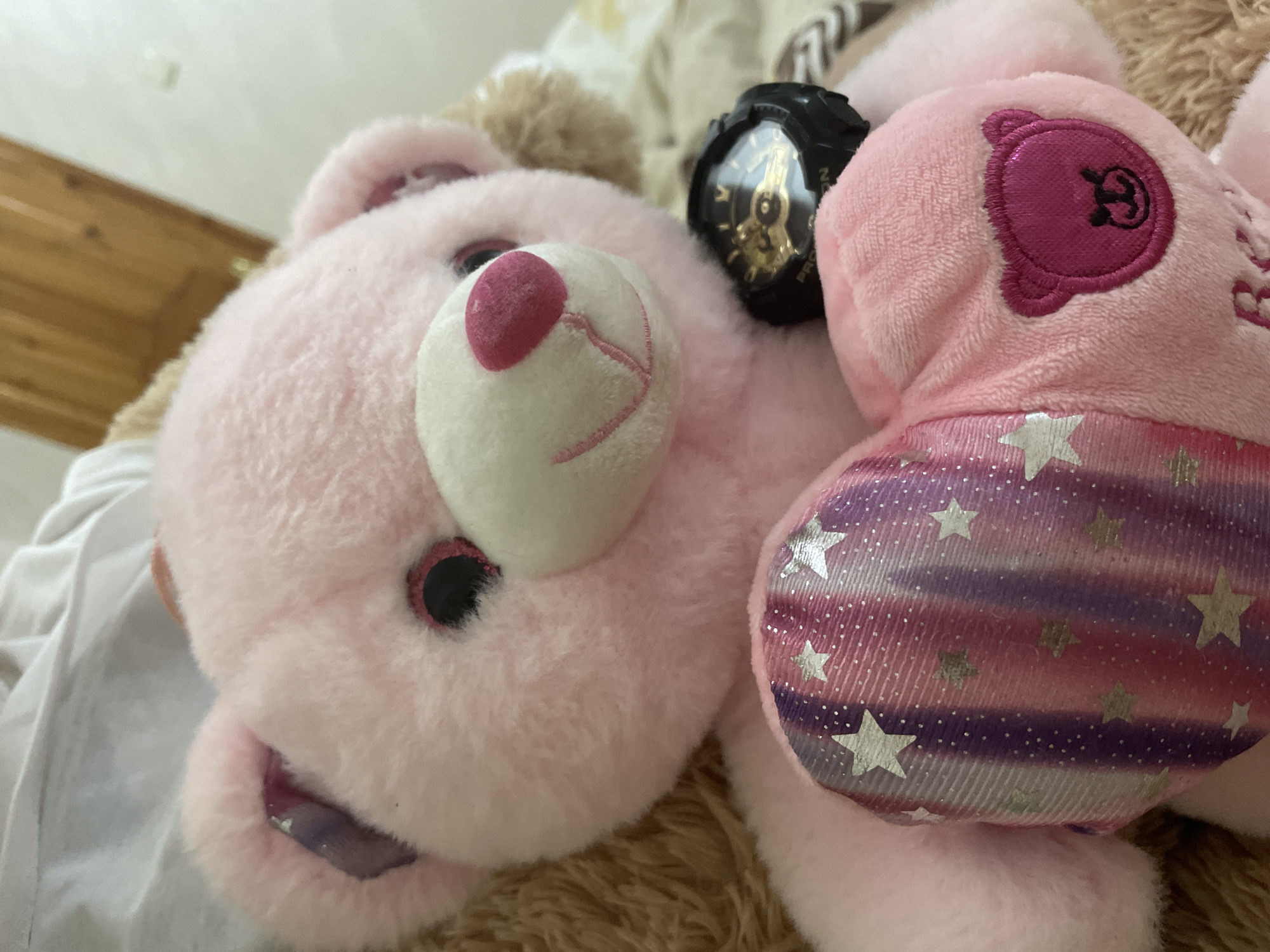 A close-up, slightly angled shot captures a pink stuffed animal, resembling a bear, resting on soft brown and white textured surfaces indoors. The toy features fluffy pink fur, round black eyes with pink glittery surrounds, a prominent white snout with a dark pink nose, and an embroidered smile. Its mouth is slightly open, revealing an iridescent, purple-blue tongue or lining.
A black digital wristwatch, displaying golden internal mechanisms through its clear dome, lies on the stuffed animal's head. The word "PROTREK" is partially visible on the watch's bezel. To the right of the main toy, another pink plush element, possibly an arm or accessory, is visible. This part includes a dark magenta circular patch embroidered with a simple bear-like face and a section patterned with horizontal stripes in pink, purple, and white, embellished with numerous silver glittery stars.
The background is softly blurred, showing glimpses of a light wooden surface and white fabric. No specific activity is depicted; the stuffed animal is stationary, creating a cozy and domestic ambiance.