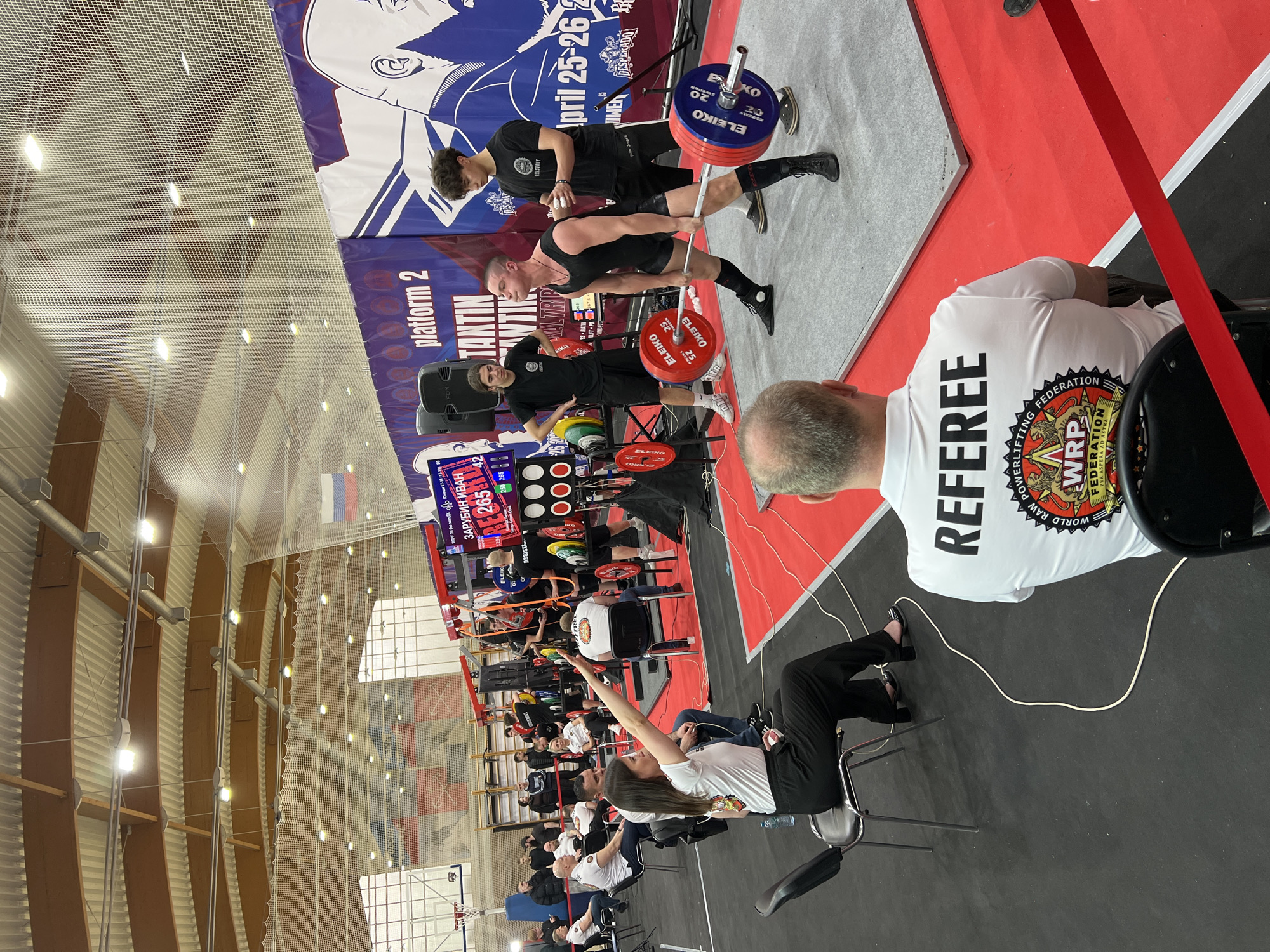 The image depicts an indoor powerlifting competition, likely in Saint Petersburg, Russia, evidenced by the Russian flag. A male powerlifter is lifting a barbell loaded with red and blue Eleiko weight plates. Two spotters stand nearby. A scoreboard displaying "ZAPYBWH HBAH RE 265" is visible, along with "platform 2" and the name "TANTIN". An official wearing a "REFEREE" shirt and the "WRPF" (World Raw Powerlifting Federation) logo is seated. Spectators sit in rows, with one woman raising her hand. The background includes banners and the architecture of a large indoor venue.