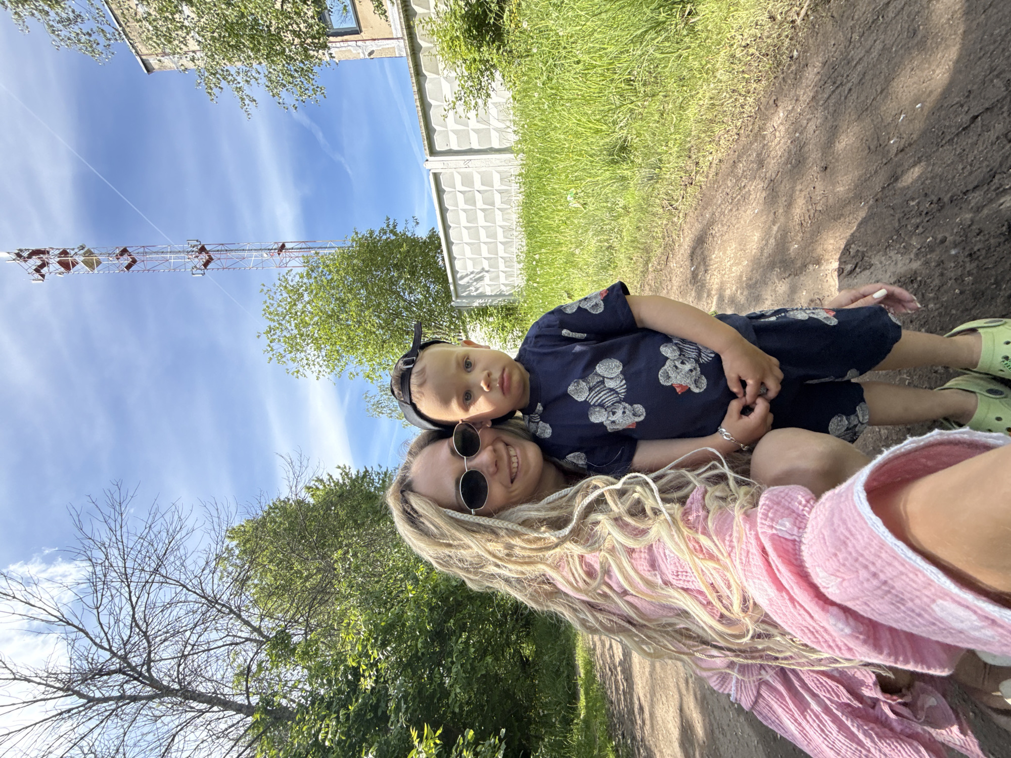 The image captures an outdoor scene in Rusanovo, Russia, featuring a woman and a child on a dirt path under a bright blue sky with scattered clouds. In the lower left foreground, a woman with long, light-colored braided hair and dark oval sunglasses crouches, smiling directly at the camera. She is wearing a light pink, textured garment. Adjacent to her on the right, a young boy stands, wearing a dark blue t-shirt adorned with a light-colored teddy bear pattern, dark blue shorts, a backward black baseball cap, and light green clogs. He looks forward with a neutral expression, his hands clasped together in front of him.
The ground consists of a brown dirt path, exhibiting tire tracks and various small debris, with shadows suggesting direct overhead sunlight. Dense green foliage, including trees and bushes, lines the left side of the path and extends into the mid-ground. On the right side, a strip of tall green grass separates the path from a white concrete fence featuring a repetitive geometric relief pattern. Beyond the fence on the far right, a light-colored, multi-story building with a visible window on an upper floor stands. A prominent red and white lattice cell tower, equipped with multiple antennae, rises vertically in the background against the sky, with a faint linear contrail visible to its left.