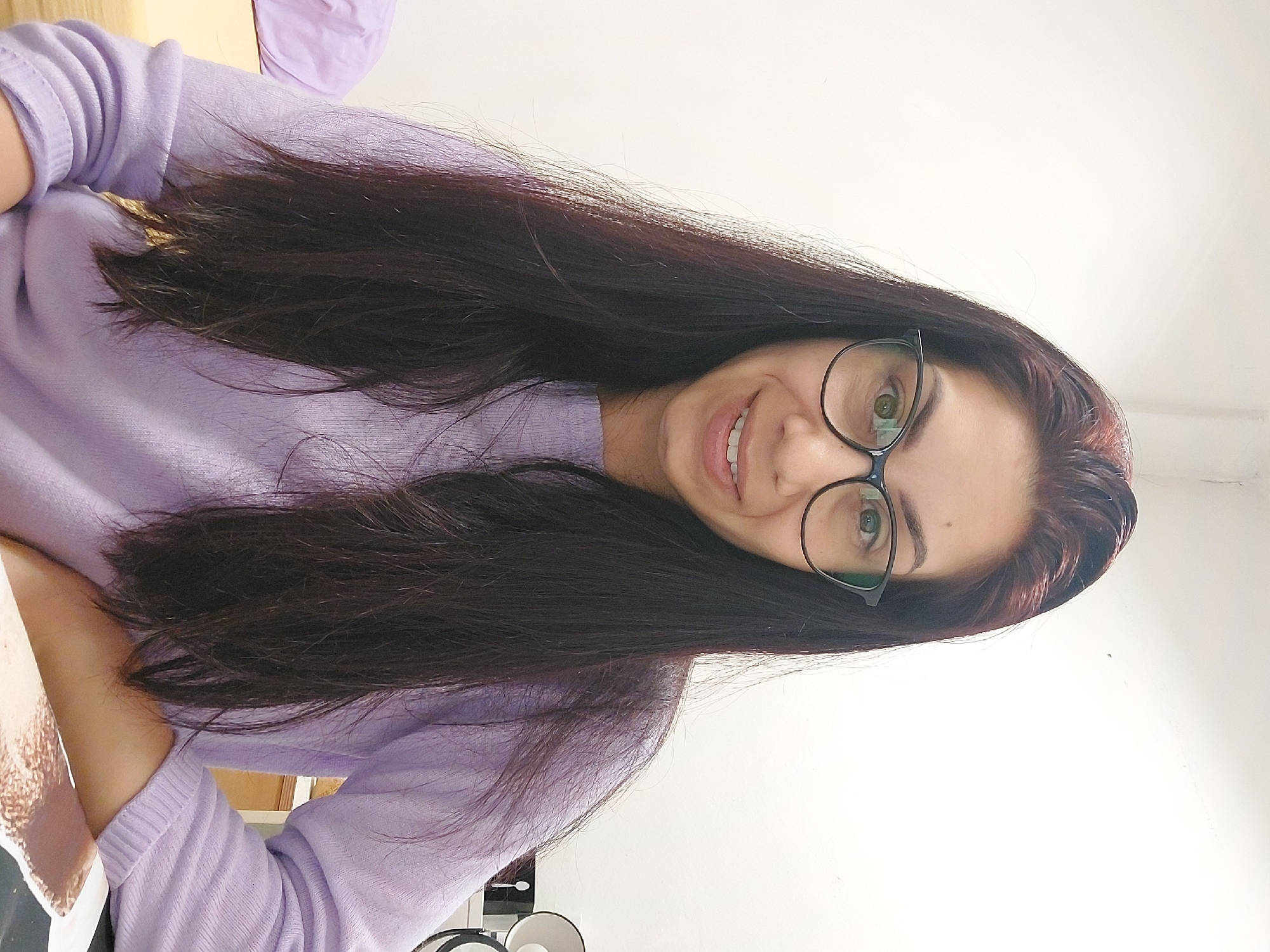 A woman with long, dark hair and glasses is looking at the camera. She is wearing a light purple sweater. She is sitting in front of a white wall. There is a desk to her right, with a lamp and some papers on it. The woman is smiling. The image is taken in Landshut, Germany.