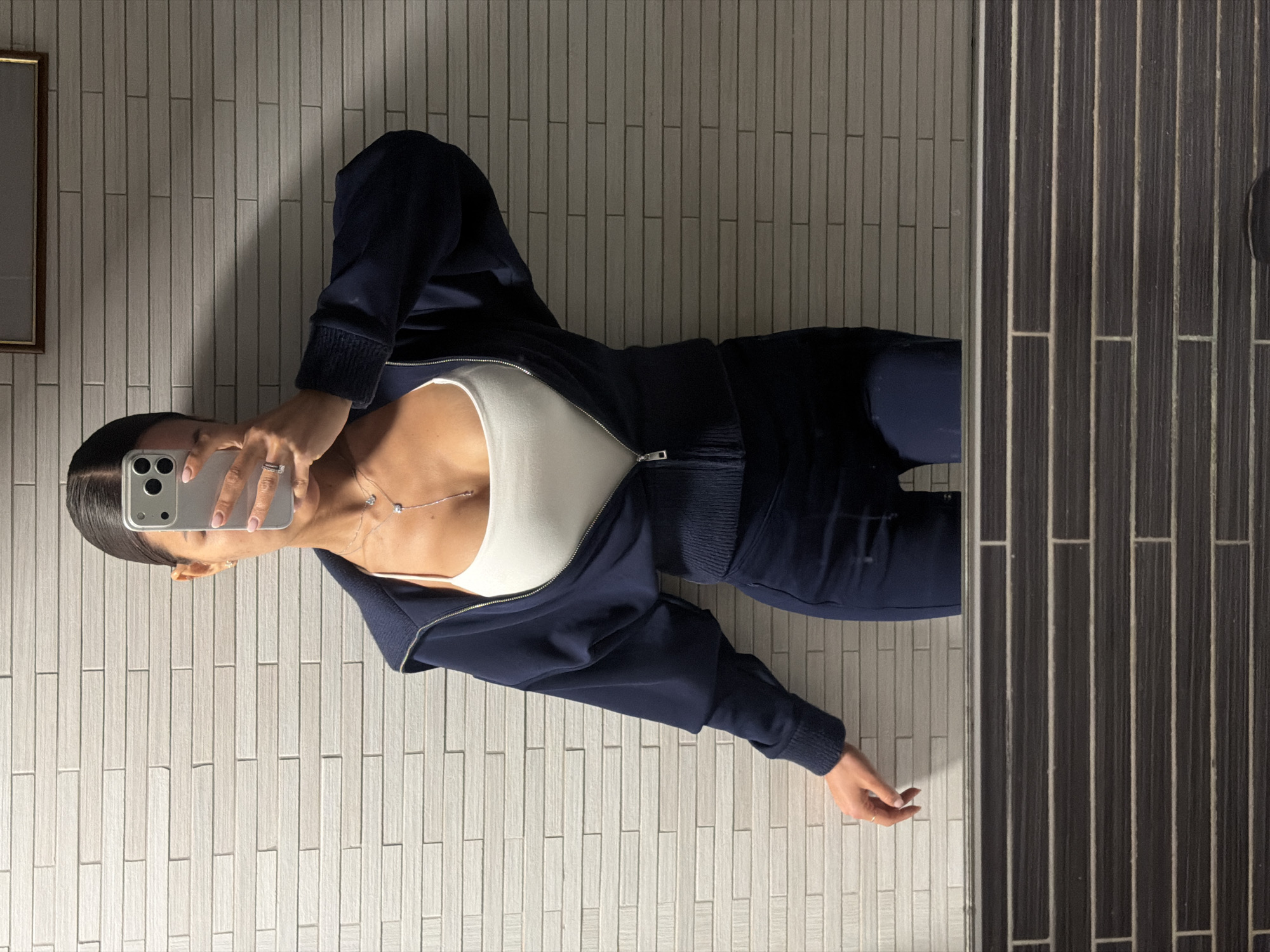 A young woman stands in front of a wall of light-colored, narrow, rectangular tiles, taking a selfie with a silver mobile phone. She wears a white top and a dark blue zip-up jacket with matching pants. Her dark hair is pulled back. To the right of the frame is a section of black and gray vertically-aligned tiles, possibly a shower or another wall surface. No information suggests that this is in Krasnoyarsk, Russia.