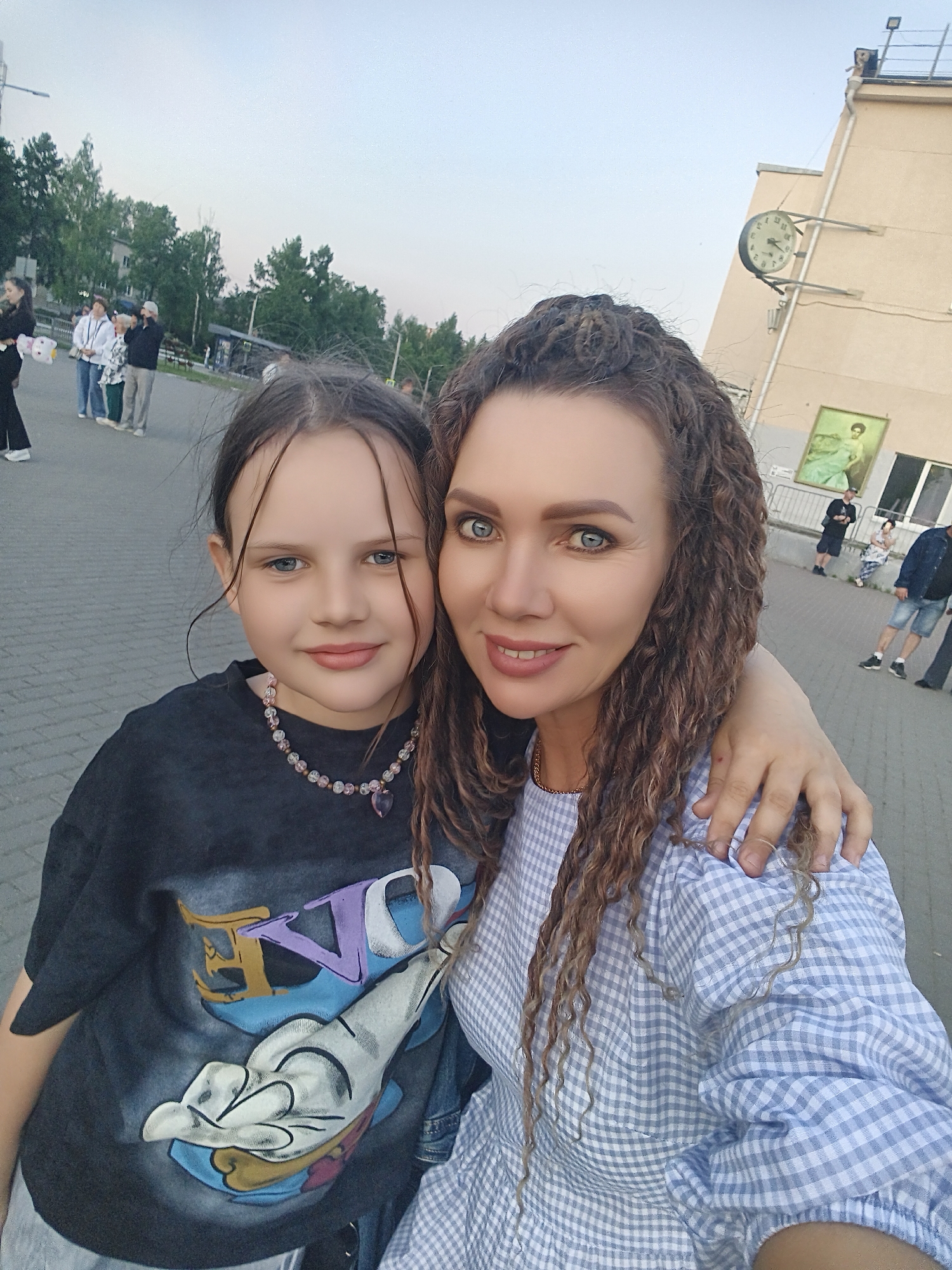 A medium-angle eye-level full shot depicts a woman and a girl posing for a selfie outdoors in Unknown, Unknown. The woman is on the right, has fair skin, curly brown hair pulled back, and blue eyes. She wears a light blue and white gingham patterned dress. Her left arm is around the girl's shoulders. The girl on the left has fair skin, light brown hair pulled back, and light blue eyes. She wears a black graphic T-shirt and a beaded necklace with a heart pendant.
In the background, a city environment is visible. Behind them is a paved area where a few people are walking or standing, including an identifiable clock on the side of a light-colored building, and green trees. The light suggests it's either early morning or late afternoon.