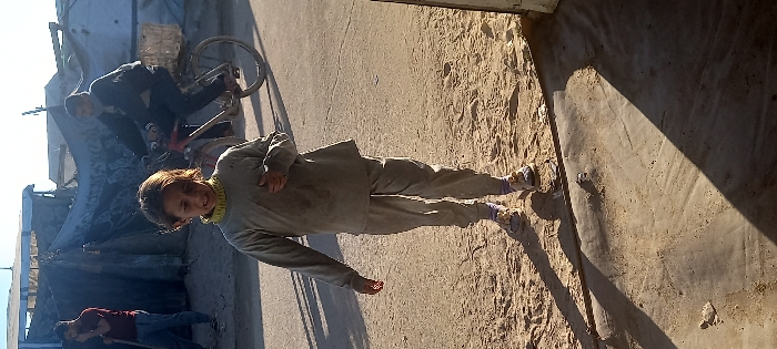 The image, oriented correctly, depicts an outdoor scene in what appears to be an informal settlement or rural area, under bright sunlight.
In the foreground, a young person, likely a girl, stands centrally, looking directly at the viewer with a slight smile or smirk. She has light brown hair and is wearing a light grey or beige long-sleeved shirt with a distinctive yellow-green ribbed collar, and matching trousers. Her shoes are light-colored, and purple socks are visible. Her left arm is slightly bent, hand open, while her right hand is near her chest. She stands on a surface that transitions from packed dirt to a more paved or hardened light-colored ground. Long, distinct shadows are cast towards the left, indicating strong sunlight from the right, suggesting either morning or late afternoon.
In the midground, behind the central figure and slightly to her left, another young person, possibly a boy, is visible. He wears dark clothing and is positioned over a bicycle, appearing to be standing next to it or preparing to ride.
The background is dominated by what appear to be makeshift structures or shelters, constructed from dark blue, grey, and black tarpaulins or fabric stretched over frames. These structures suggest a humble living environment. To the far left in the background, another person is partially visible, wearing a red shirt and blue jeans, bending over near the base of one of these shelters. The sky visible in the upper left corner is bright and clear blue.
The overall impression is one of children playing or going about daily life in a modest, outdoor setting with rudimentary dwellings. No discernible text is visible in the image.