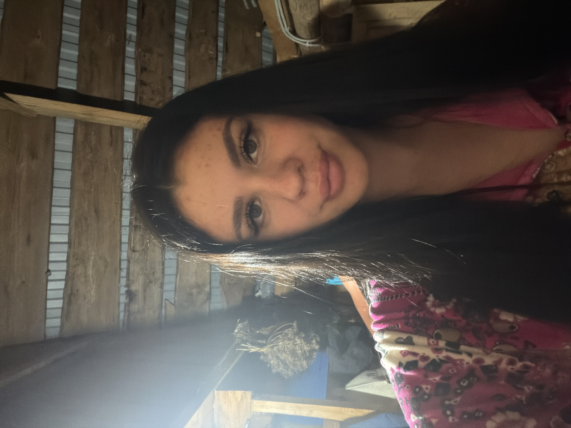 The image shows a young woman with long, dark hair and prominent makeup taking a selfie in what appears to be an attic or barn. The background consists of wooden beams and corrugated metal sheeting, suggesting a rustic or unfinished interior. A bundle of dried plants hangs from a beam on the left side of the frame, and various objects are visible in the dimly lit background, including what might be storage containers or tools. The woman is wearing a pink shirt with a patterned design. The lighting is dim and uneven, with a strong light source from the left creating a lens flare effect. The specific location in the city of Фоки, Russia, is not discernible from the image itself.