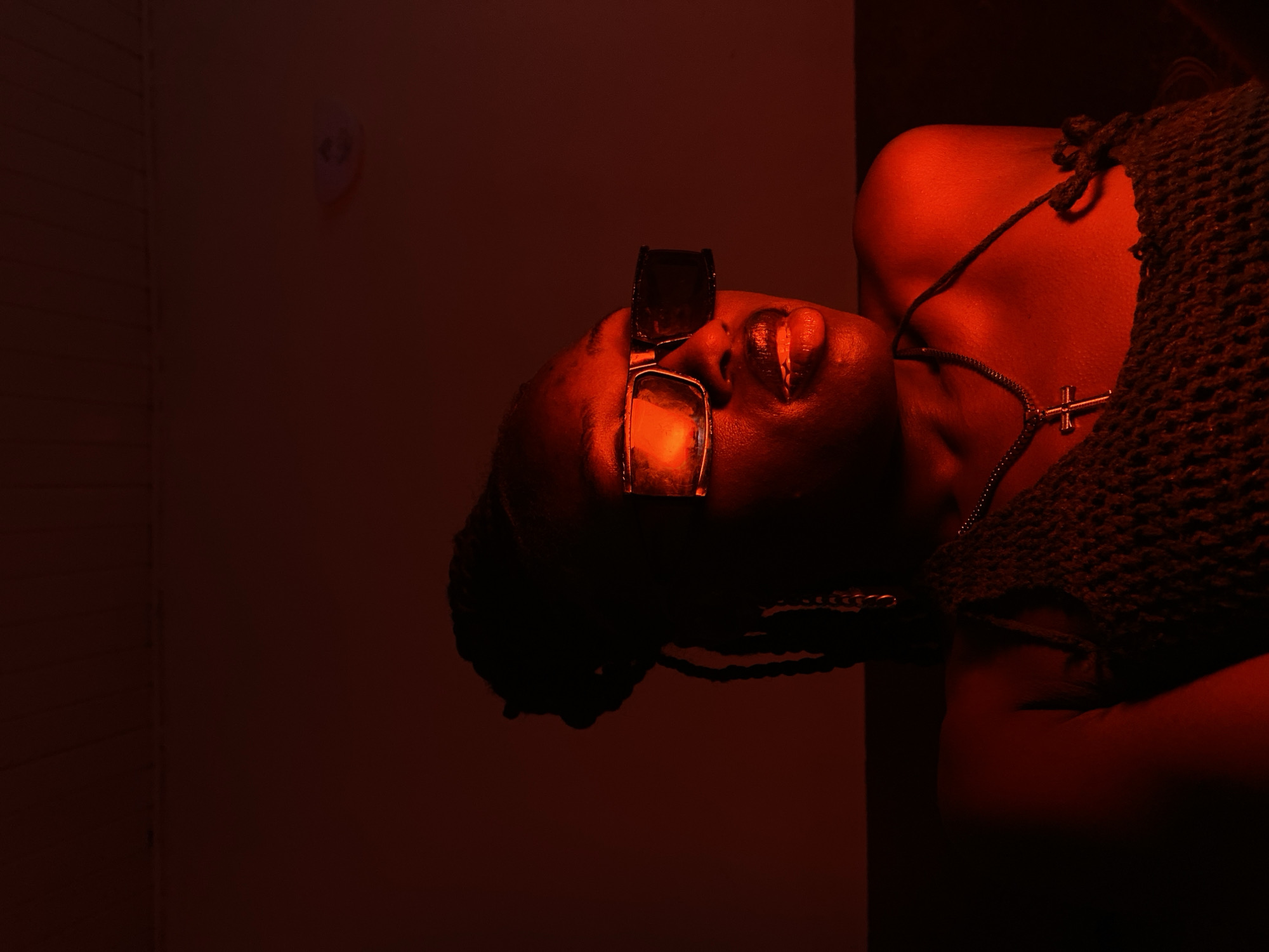 A medium shot captures a young Black woman from the chest up, bathed in a strong red light. She wears large, dark-rimmed sunglasses with thick frames that obscure her eyes. Her lips are parted, revealing her tongue slightly, and her skin appears to be glistening. She has dark braided hair that hangs down her shoulders, and a silver cross pendant hangs from a chain around her neck, resting on a dark, textured top. The background is a plain, reddish-brown wall, with some subtle texture suggesting it might be plaster or painted concrete. There is a circular object mounted on the wall to the upper right of the frame, possibly a smoke detector or light fixture. The overall ambiance is moody and artificial due to the intense red lighting. The scene is set indoors.
