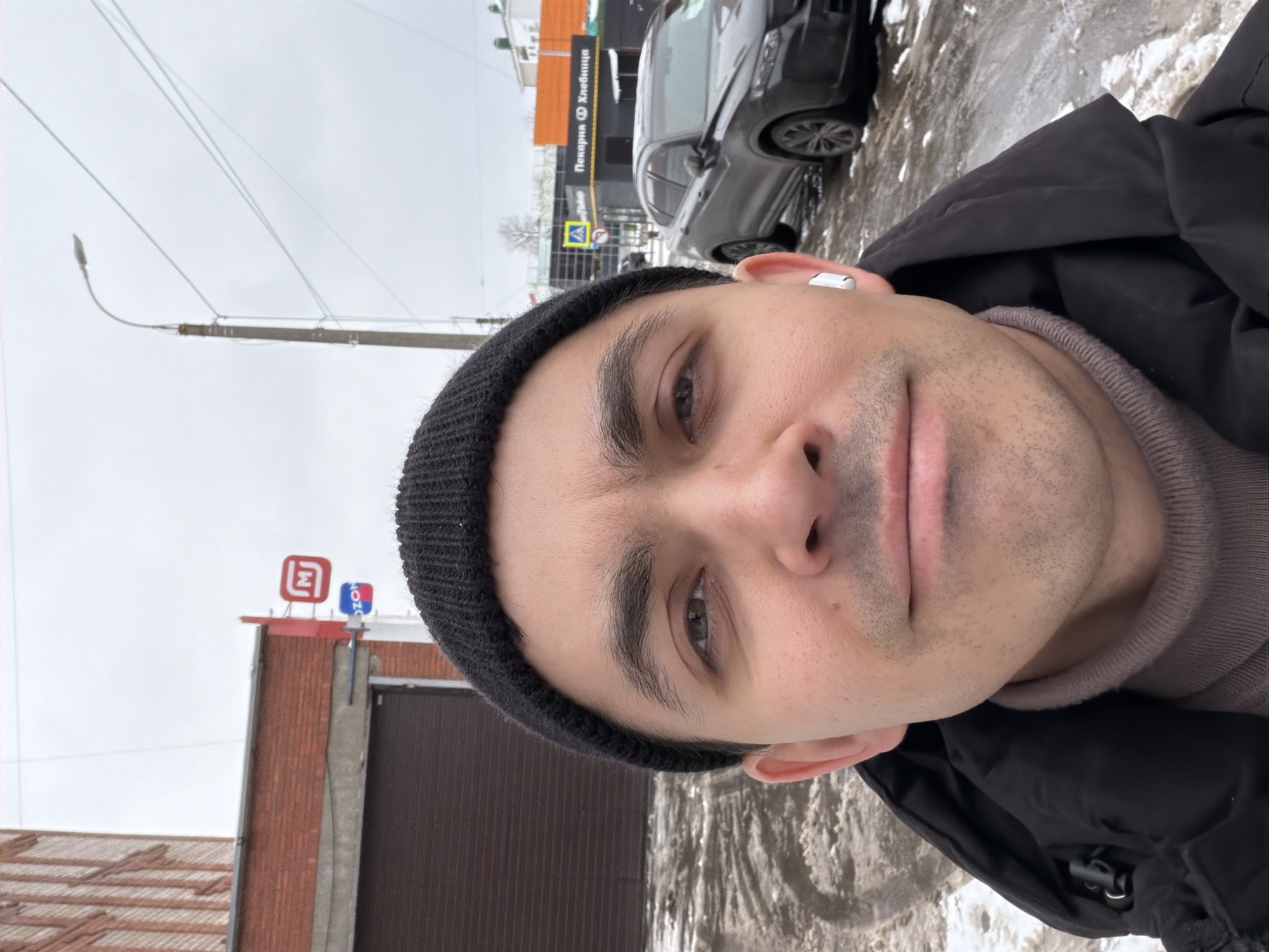 The image depicts a selfie of a man wearing a black beanie, a beige turtleneck sweater, and a black jacket. He has short dark hair, dark eyebrows, and light skin. He has a wireless earbud in his right ear.
In the background, there's a street scene with buildings, a car, and some snow on the ground. The building signage indicates the location is Izhevsk, Russia, with the presence of "OZON" (online retailer) and "Pekarnya Khlebnitsa" (Bakery Bread). A street lamp is visible. The weather appears overcast.