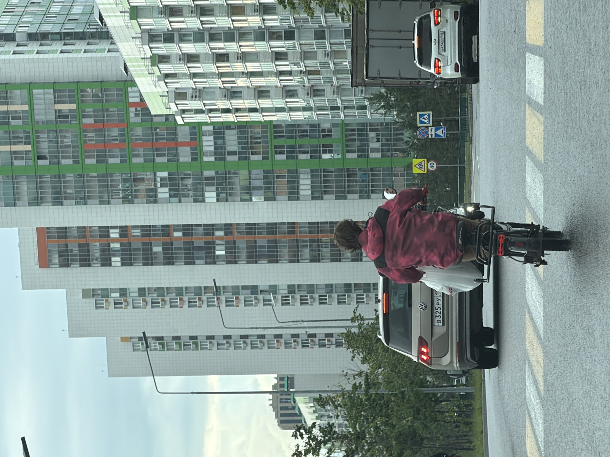 In an urban setting, a person rides a bicycle on a wet asphalt road, approaching a pedestrian crossing. The cyclist, wearing a red hooded jacket, carries a white plastic bag. In the background, several modern apartment buildings rise into a cloudy sky. The buildings feature a mix of white facades with colorful accents in green, orange, and beige, and numerous windows. Several cars and a large truck are visible on the road, with traffic lights and road signs indicating intersections and speed limits. Trees line the left side of the road. The overall scene suggests a typical day in a city with ongoing construction or development.