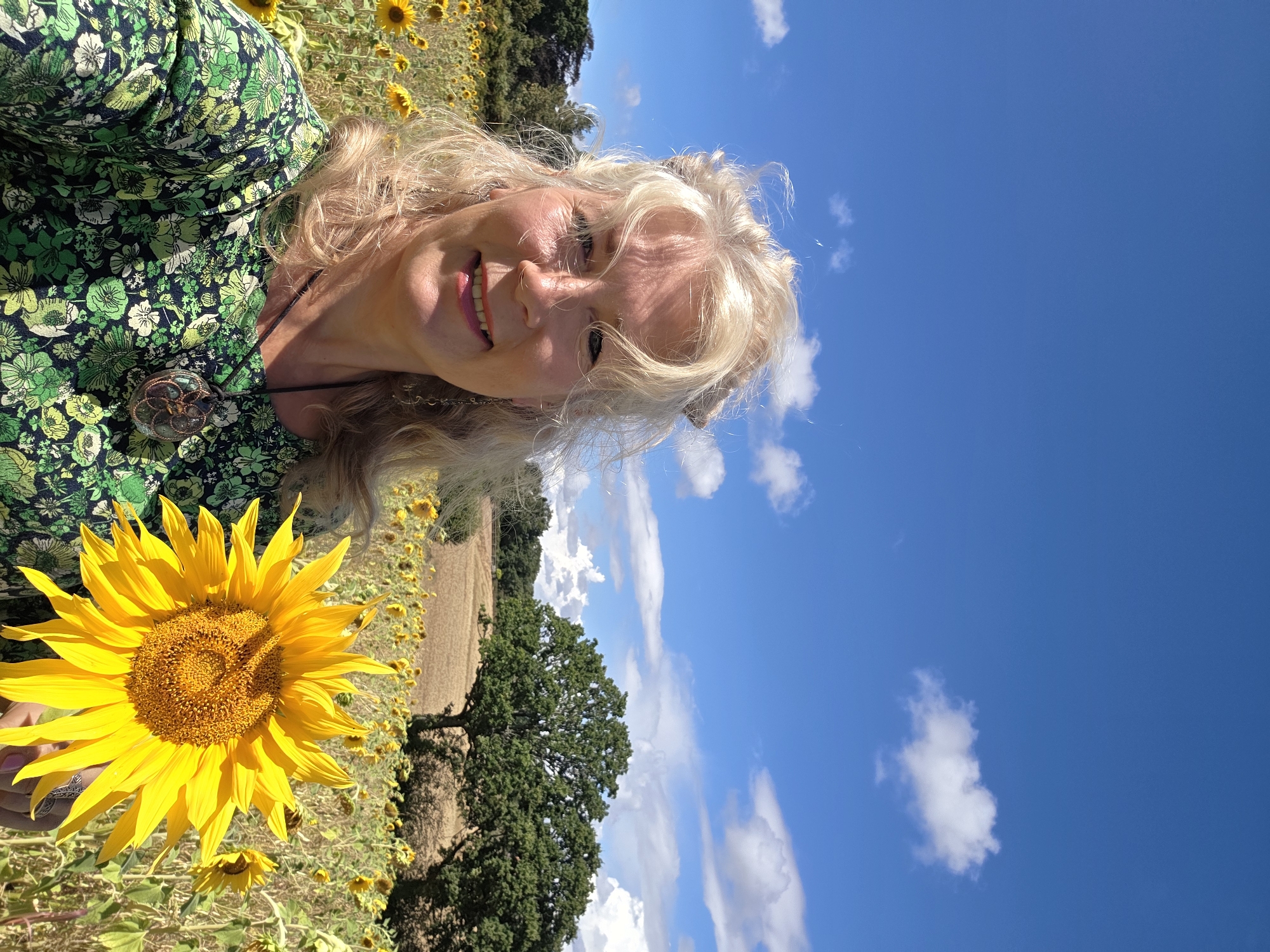 A woman with blonde hair and fair skin is standing in a sunflower field under a blue sky with white clouds. She is smiling and holding a large sunflower in her right hand. She is wearing a green floral dress and a necklace. In the background, there are more sunflowers, trees, and a harvested field. The image appears to have been taken on a sunny day. The location is the city of Rother, United Kingdom.