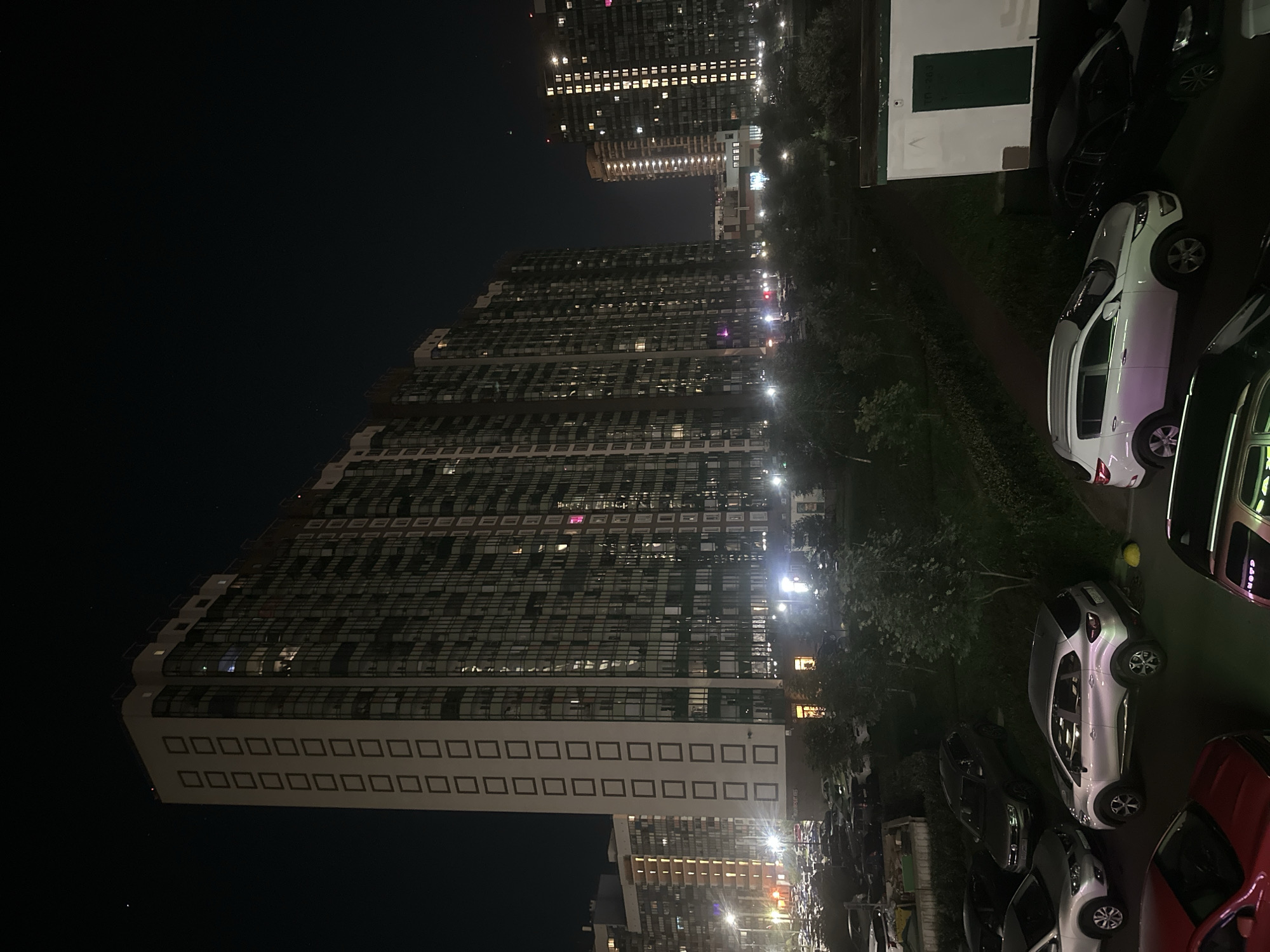 A nighttime, high-angle, exterior shot captures a modern, multi-story residential building in the city of Sinyavino, Russia. The building, a dominant feature, has a white facade adorned with a vertical grid of dark square window frames and is illuminated by lights from within. The dark sky above is dotted with a few faint stars. In the foreground, a parking lot is filled with numerous cars of various models and colors, mostly dark. Adjacent to the parking lot, there is a landscaped area with trees and bushes. To the right of the frame, a smaller, white building with a green door is visible. The overall scene depicts a residential area at night.