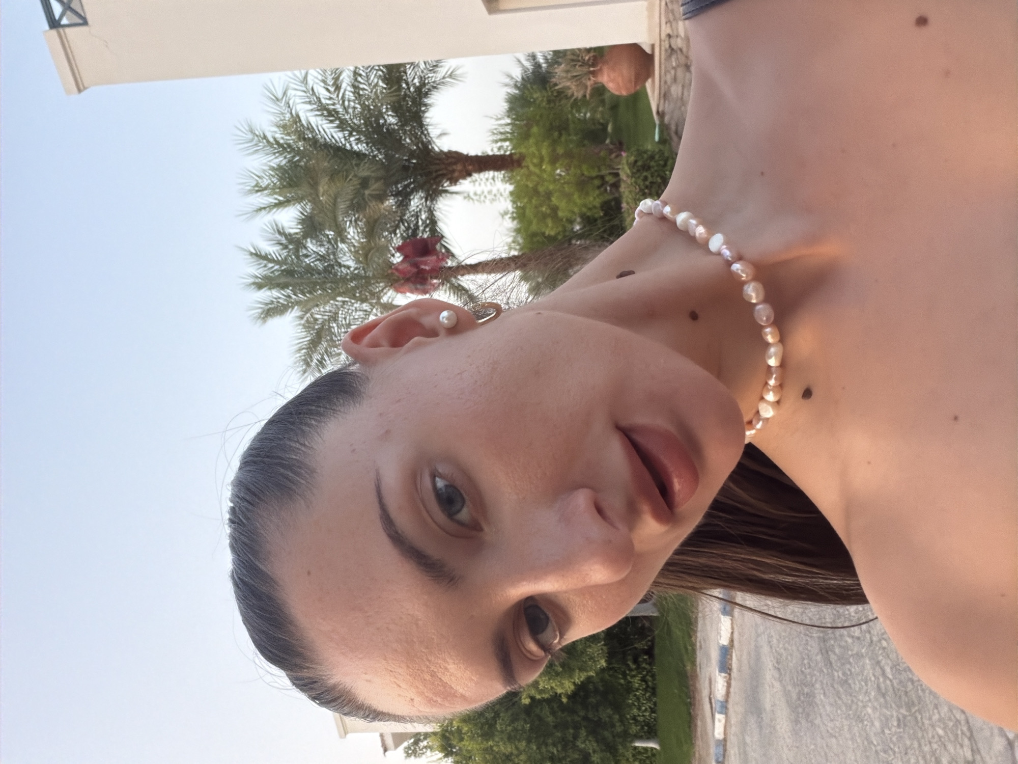 An outdoor, medium shot portrait of a young Caucasian woman with her face tilted downwards and to her right, against a backdrop of a clear sky, palm trees, and a building. She has dark hair pulled back in a ponytail, fair skin, blue eyes, and is wearing a pearl necklace and hoop earrings. The woman is positioned slightly off-center to the left, with her right shoulder and collarbone visible. The environment appears to be a resort or residential area in Sharm El Sheikh, Egypt, characterized by lush greenery, palm trees, and white architectural elements. The lighting suggests it is either early morning or late afternoon, with a warm, soft glow.
