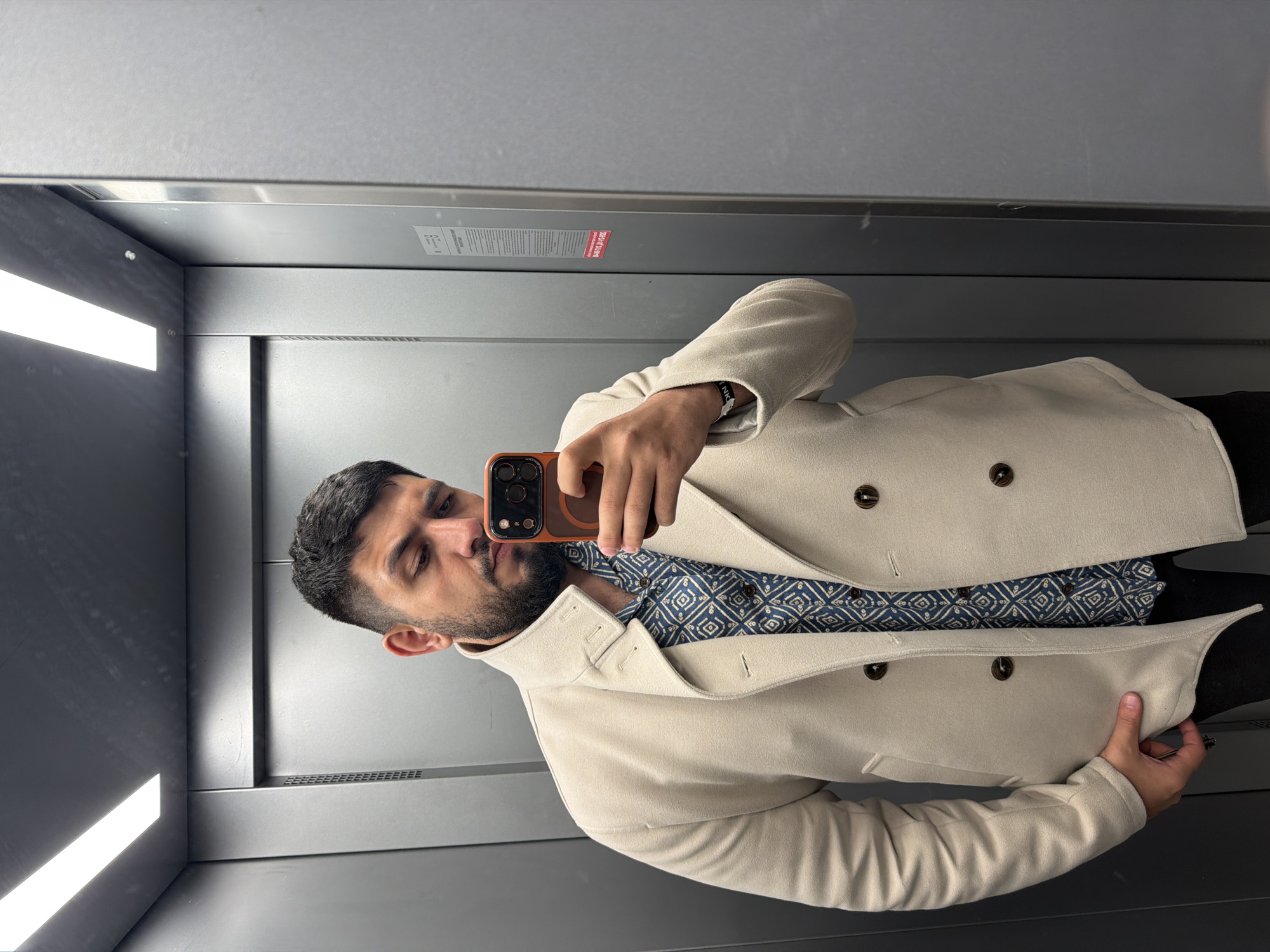 A man with dark hair and a beard is taking a selfie in an elevator. He is wearing a beige overcoat over a patterned blue shirt. The elevator has gray metal walls and fluorescent lighting on the ceiling. There is a small red sticker near the top of the back wall. The location in the city of Krasnoyarsk, Russia is not discernible from the image.