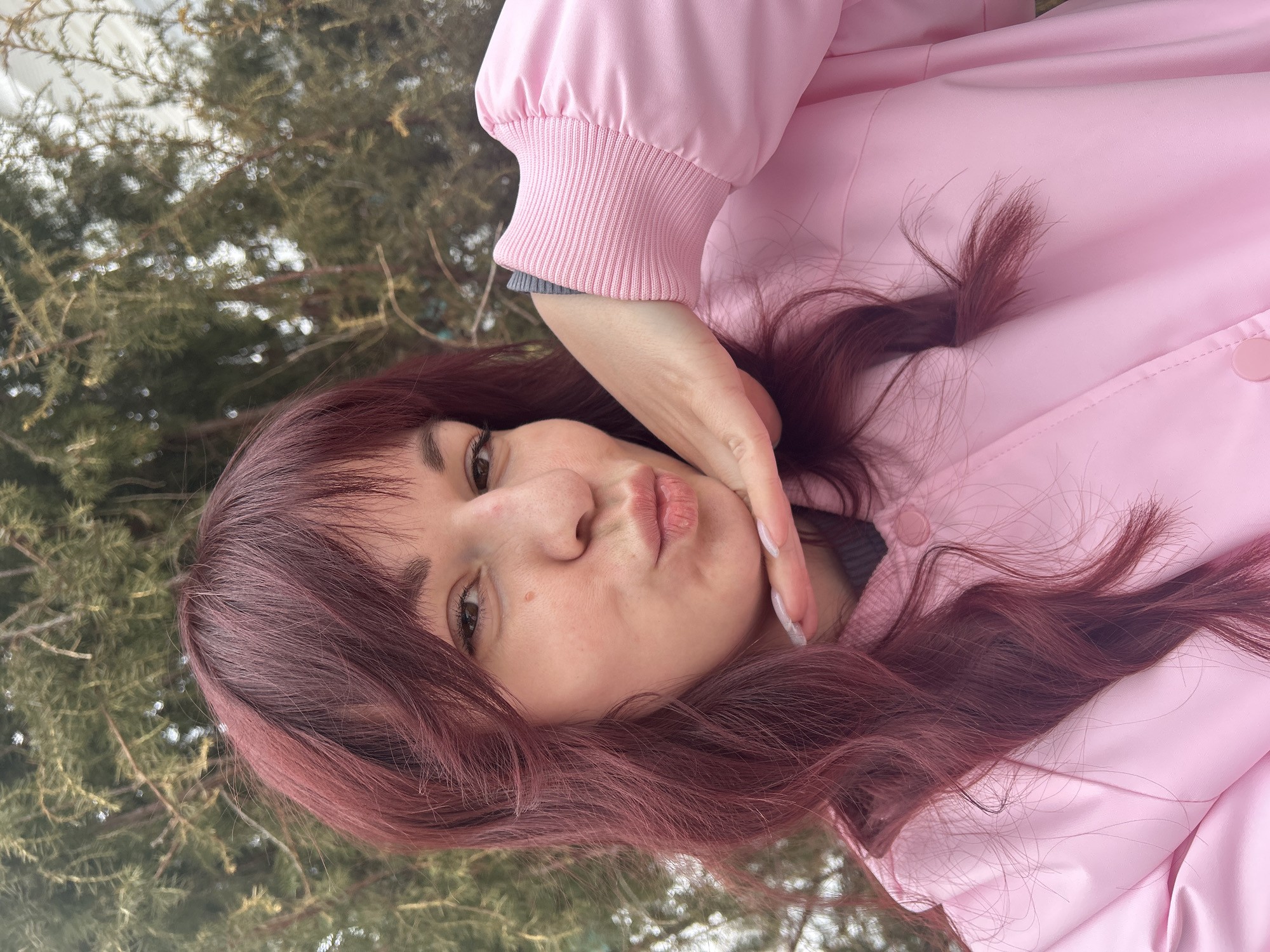 A woman with dark red, wavy hair and brown eyes poses for a selfie outdoors in Фоки, Russia. She is wearing a light pink jacket and a grey sweater underneath. Her hand is touching her face, and she is making a kissing face. The background includes blurry greenery, likely trees.