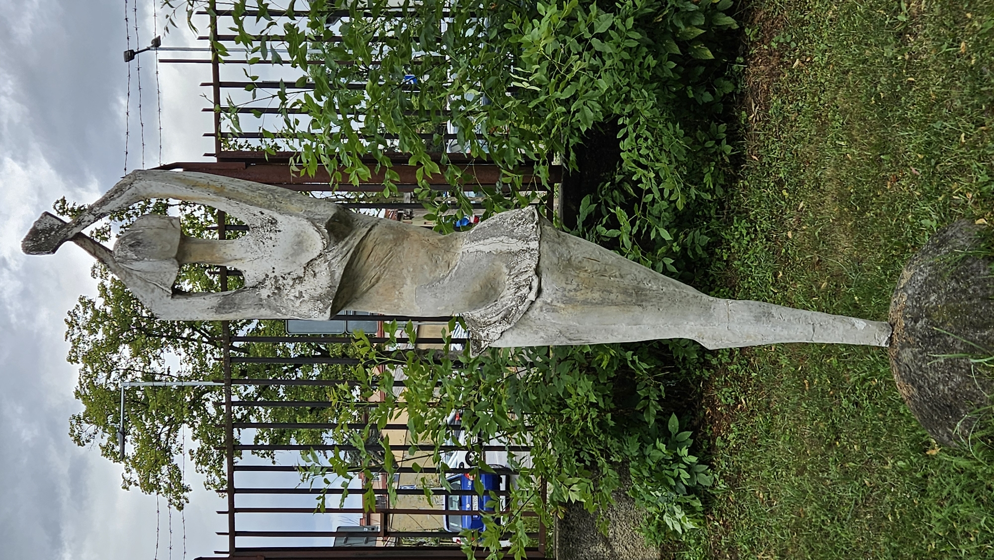 A concrete statue of a stylized human figure stands on a grassy area in front of a metal fence. The figure, which is abstract and appears to be female, is reaching upwards with both arms. The statue is weathered and shows signs of age. Behind the statue and fence, trees and buildings are visible, along with a few cars on a street. The sky is overcast. This statue is located in Prešov, Slovakia.
