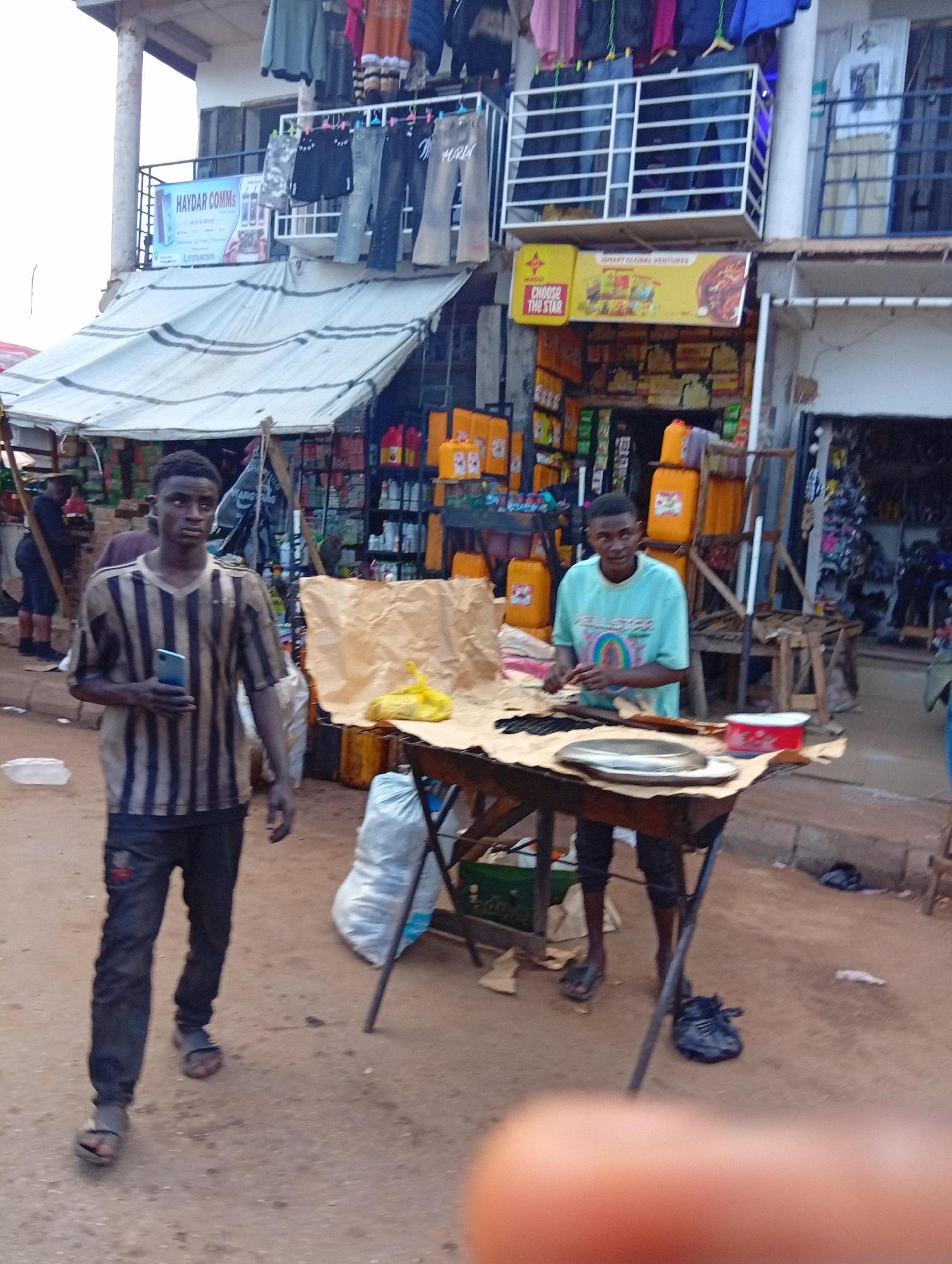 In the city of Bukuru, Nigeria, a street scene unfolds outside a shop. Two young men are the primary subjects. The man on the left, wearing a striped t-shirt and dark pants, holds a smartphone in his right hand and walks towards the viewer. He is wearing sandals. The man on the right, dressed in a light blue t-shirt and dark pants, stands behind a wooden table and appears to be preparing something on a brown paper covering. He is also wearing sandals. The table is laden with a large metal plate, what looks like a red tin, and some black items. Behind the man on the right, the shop is stocked with various goods, including yellow containers, bottles, and other items, suggesting it's a retail establishment. In the background, more shops are visible, along with clothing items displayed for sale. The ground is unpaved and dusty.