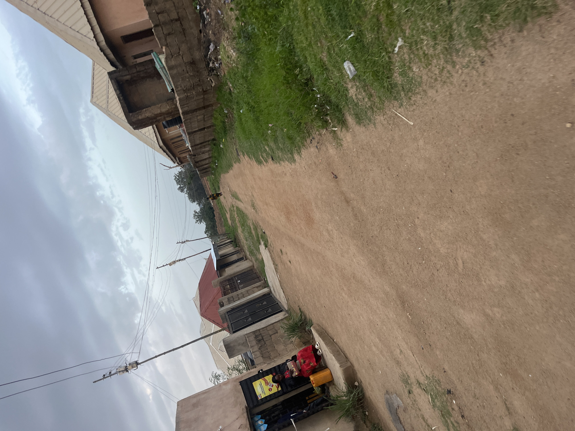 The image depicts a street scene in Jos, Nigeria, under an overcast sky. The street is unpaved and appears to be made of dirt. On the left side of the street, there is a small building with an open storefront. A woman is seated on a step in front of the store, wearing a red dress and white sandals, and holding a box. Beside her sits a yellow container. A person is visible walking further down the street in the distance. The right side of the street features a wall made of bricks and concrete, with overgrown grass and scattered debris along its base. Several buildings with corrugated metal roofs are visible along the street, some appearing to be residential. Utility poles with wires are present, indicating the presence of electricity infrastructure. The overall impression is one of a developing urban or semi-urban environment.