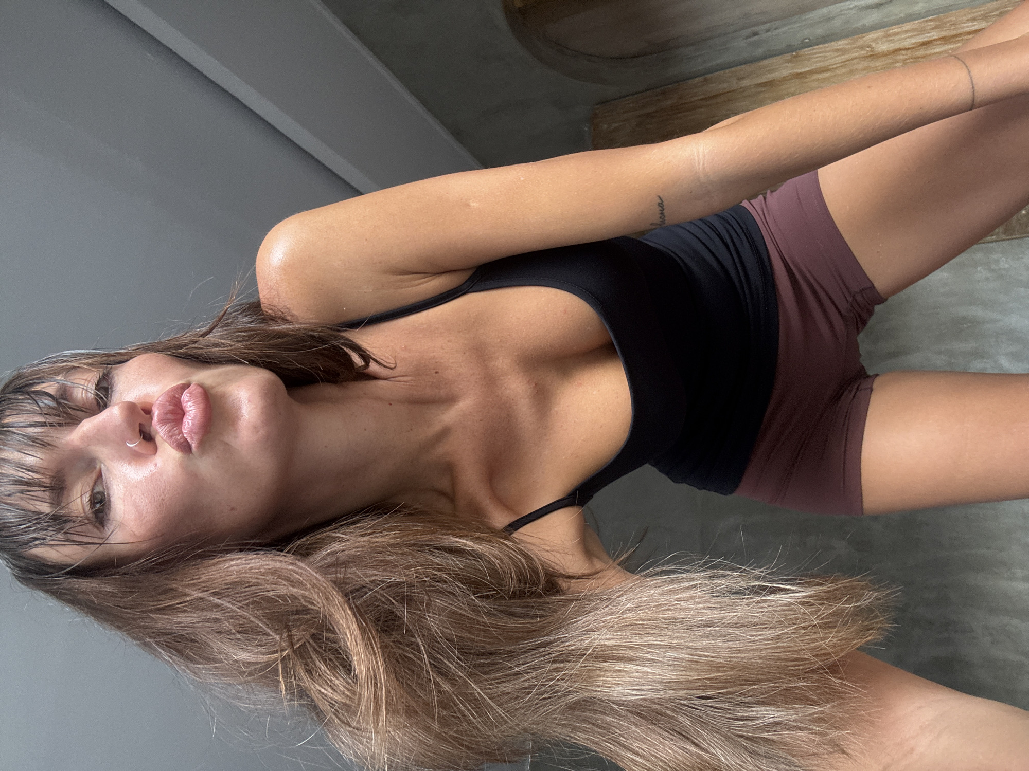 A woman with light brown hair and a nose ring is lying on a gray surface. She is wearing a black tank top and brown shorts. Her hair is wet and falling across her face. The location appears to be indoors, in North Kuta, Indonesia.