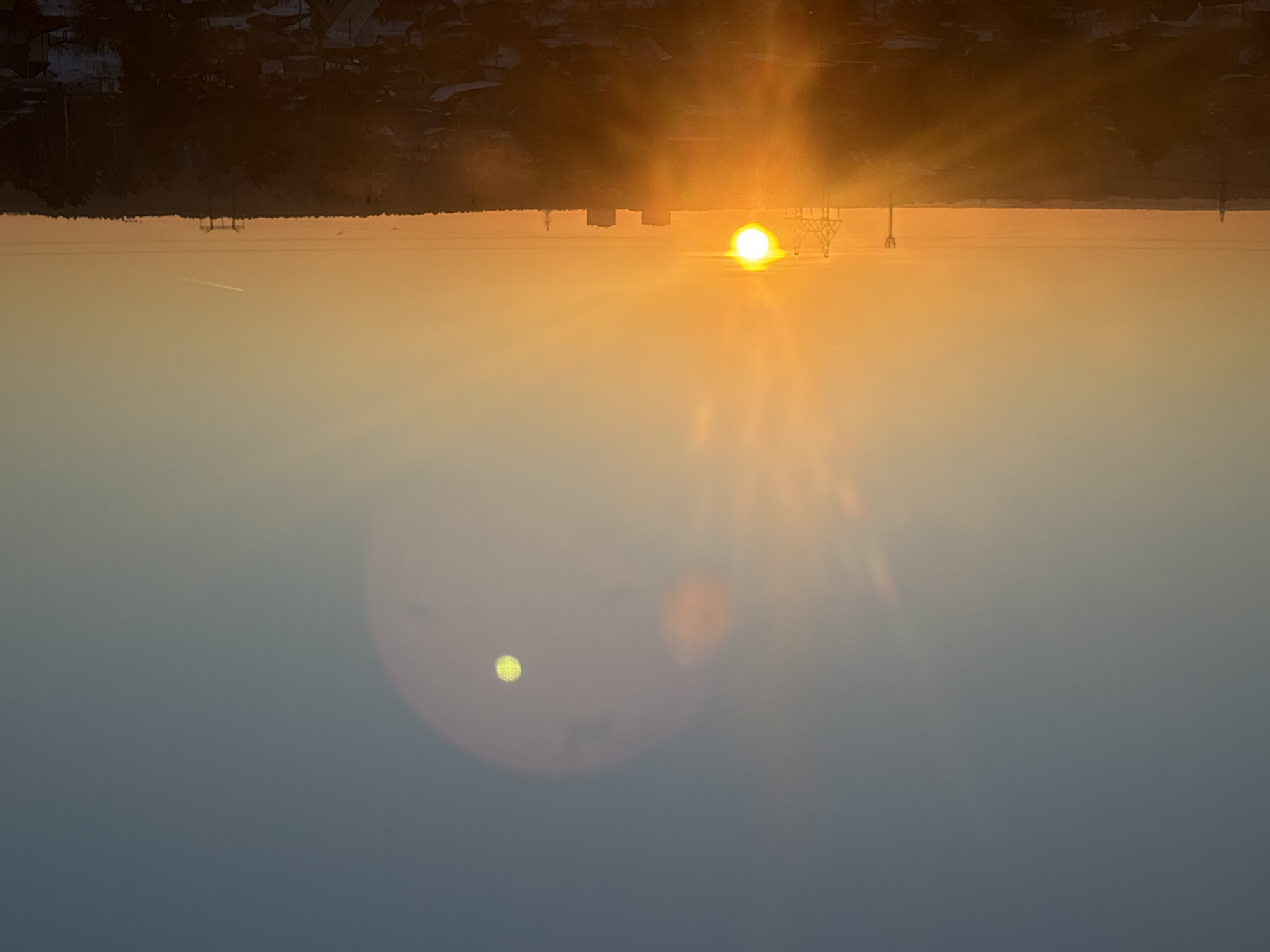 An elevated, wide-angle photograph depicts a sunset or sunrise over the city of Chaikovsky, Russia. The sky transitions from light blue at the top to vibrant orange and yellow at the horizon, with the sun positioned low and slightly left of center, emitting strong light rays and causing prominent lens flares, including a distinct green-yellow circular artifact in the upper right sky. A faint contrail is visible in the upper right quadrant. The horizon is marked by a dark, dense tree line, above which several industrial and urban structures are silhouetted: a tall transmission tower on the left, four interconnected industrial structures (possibly cranes) further right, two distinct dark rectangular high-rise buildings, and another slender industrial tower on the far right. The midground and foreground reveal a sprawling residential area, largely in shadow, characterized by numerous houses with dark roofs and bare trees. Patches of snow are visible on the ground and rooftops, indicating a cold weather season. No people or active interactions are visible.