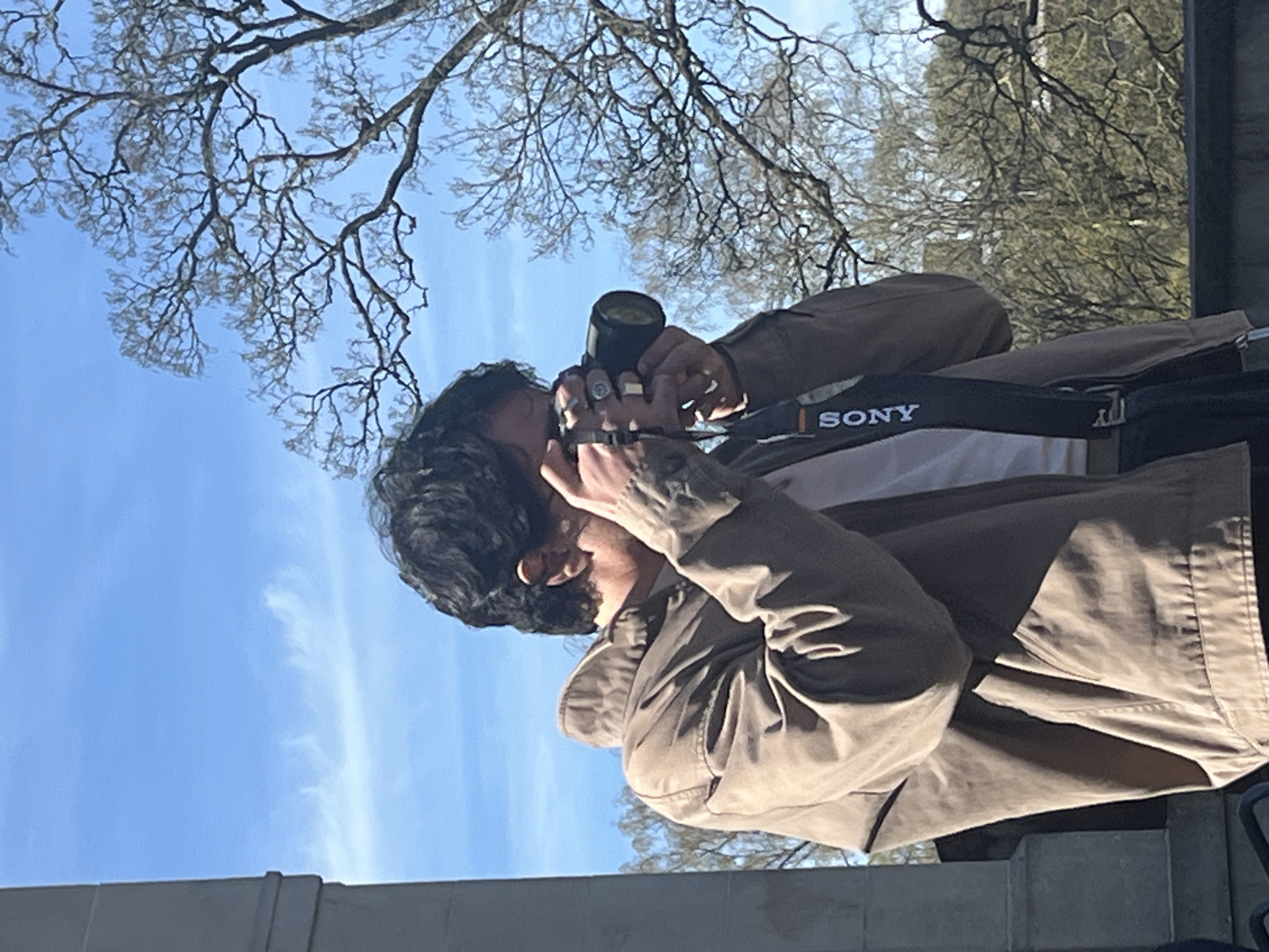 A person with dark curly hair wearing a khaki-colored jacket and a white shirt is holding a black Sony camera up to their face as if taking a photograph. The person has multiple rings on their fingers and a tattoo on their hand. The camera has a black strap with the word "SONY" on it in white letters. Behind the person is a blue sky with a few wispy clouds, and bare tree branches reach into the sky. The bottom of the image shows a concrete structure, possibly a wall or ledge. The image seems to be taken at a low angle, looking up at the person and the sky. Without additional information or contextual clues, it's impossible to confirm that the location of the image is Luxembourg City.