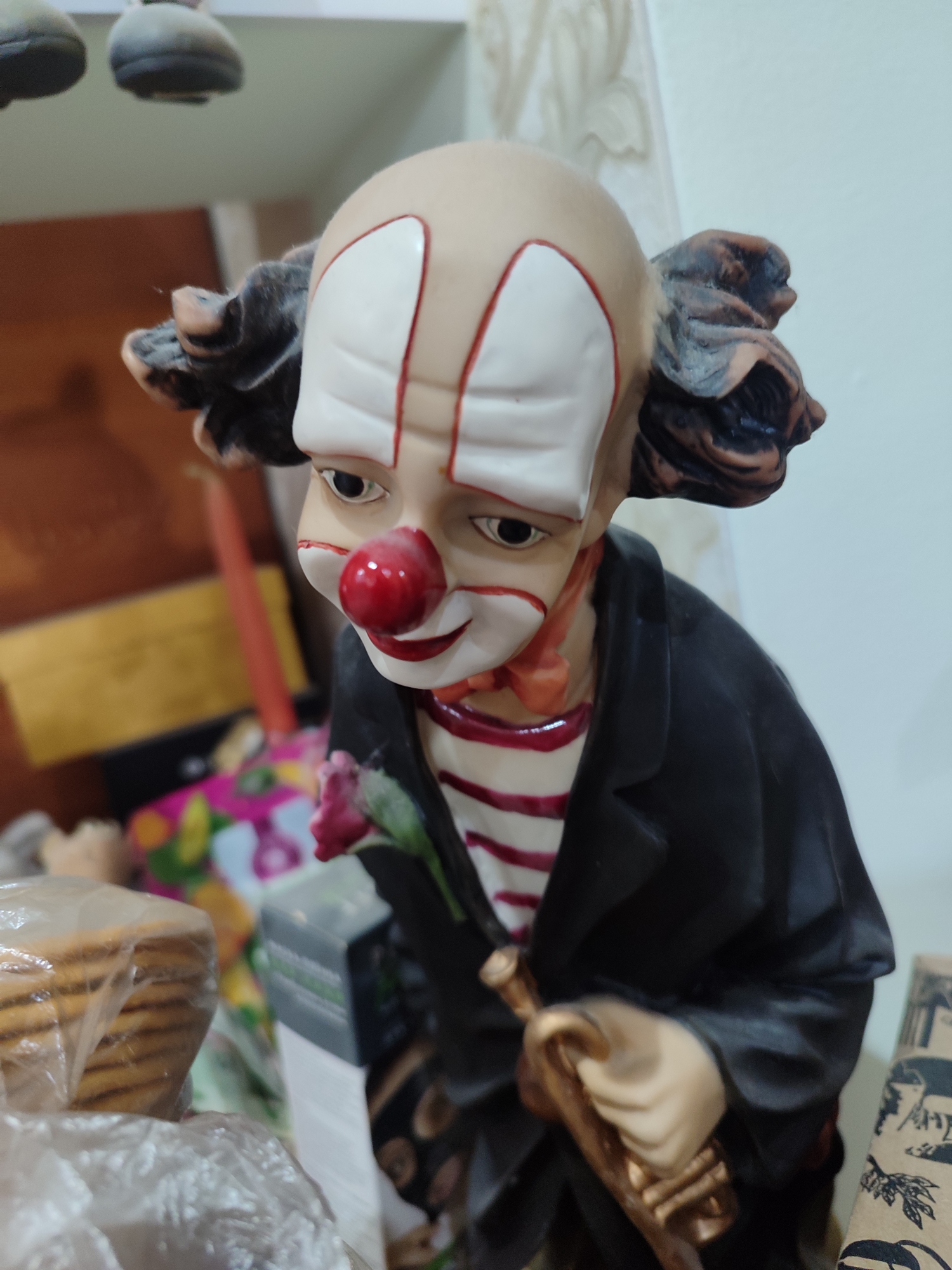 A close-up, eye-level shot focuses on a ceramic figurine of a sad-looking clown. The clown is bald, with painted white makeup on its forehead and cheeks, outlined in red. Its nose is a bright red sphere, and it has black eyes and a downturned mouth. The clown is wearing a red and white striped shirt under a black jacket. It holds a light pink rose in its left hand and a golden trumpet in its right. The background is blurred, but suggests an indoor setting with shelves and various items, including stacks of cookies in plastic packaging and a colorful floral-patterned object. The lighting appears to be artificial, and there is no indication of weather conditions. The emotional tone conveyed by the clown is somberness. There is no visible text in the image.