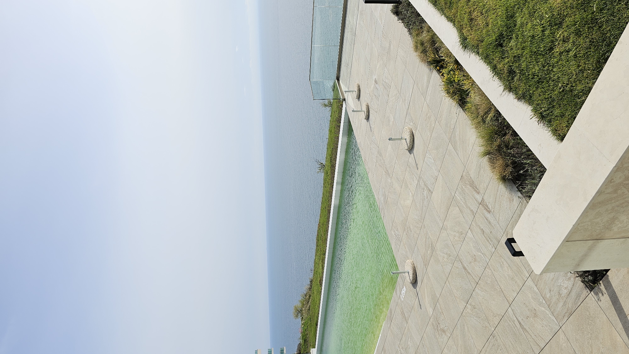 A rotated, high-angle shot shows the edge of an infinity pool extending towards the sea in Kavarna, Bulgaria. To the left, the pale sky meets the calm, blue sea on the horizon. The pool, filled with green water, runs along the right edge, bordered by a concrete ledge and green foliage along the shoreline. Parallel to the pool is a stone-tiled patio area. At regular intervals, round, stone-like features with metal protrusions are positioned on the patio. Further right, manicured grass is separated by stone borders, extending towards the upper right corner. The foreground reveals architectural elements with cream-colored stone slabs and a black light fixture. The image is mostly clear, with slight blurring from the rotation.
