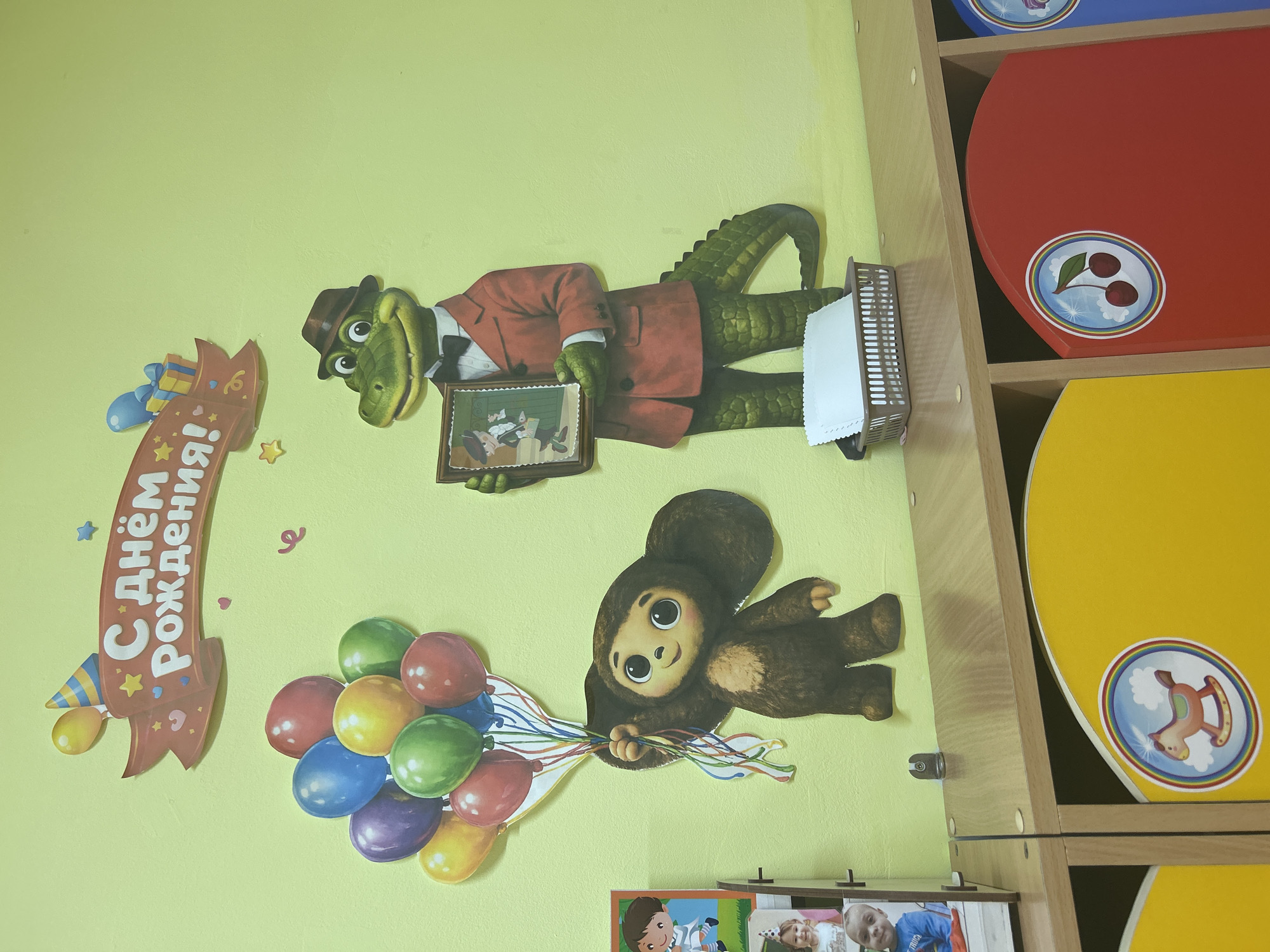 This media file shows a section of a wall decorated with cutouts. On the left, a banner with Cyrillic text reads "С Днём Рождения!" (Happy Birthday!). Beside the banner, a cartoon alligator wearing a red suit and hat is depicted holding a framed picture of other cartoon characters. Below the alligator and to the right, a brown, furry cartoon character, possibly Cheburashka, is shown holding a bouquet of colorful balloons. The wall is painted a light yellow. To the right of the wall decorations are shelves with colorful, curved surfaces in red and yellow, with circular decals featuring children's motifs like cherries and a rocking horse. In the bottom right corner, there are framed photographs of children. The overall scene suggests a child's birthday party or a decorated children's room in Gelendzhik, Russia. The bright colors and cartoon characters evoke a festive and cheerful atmosphere.
