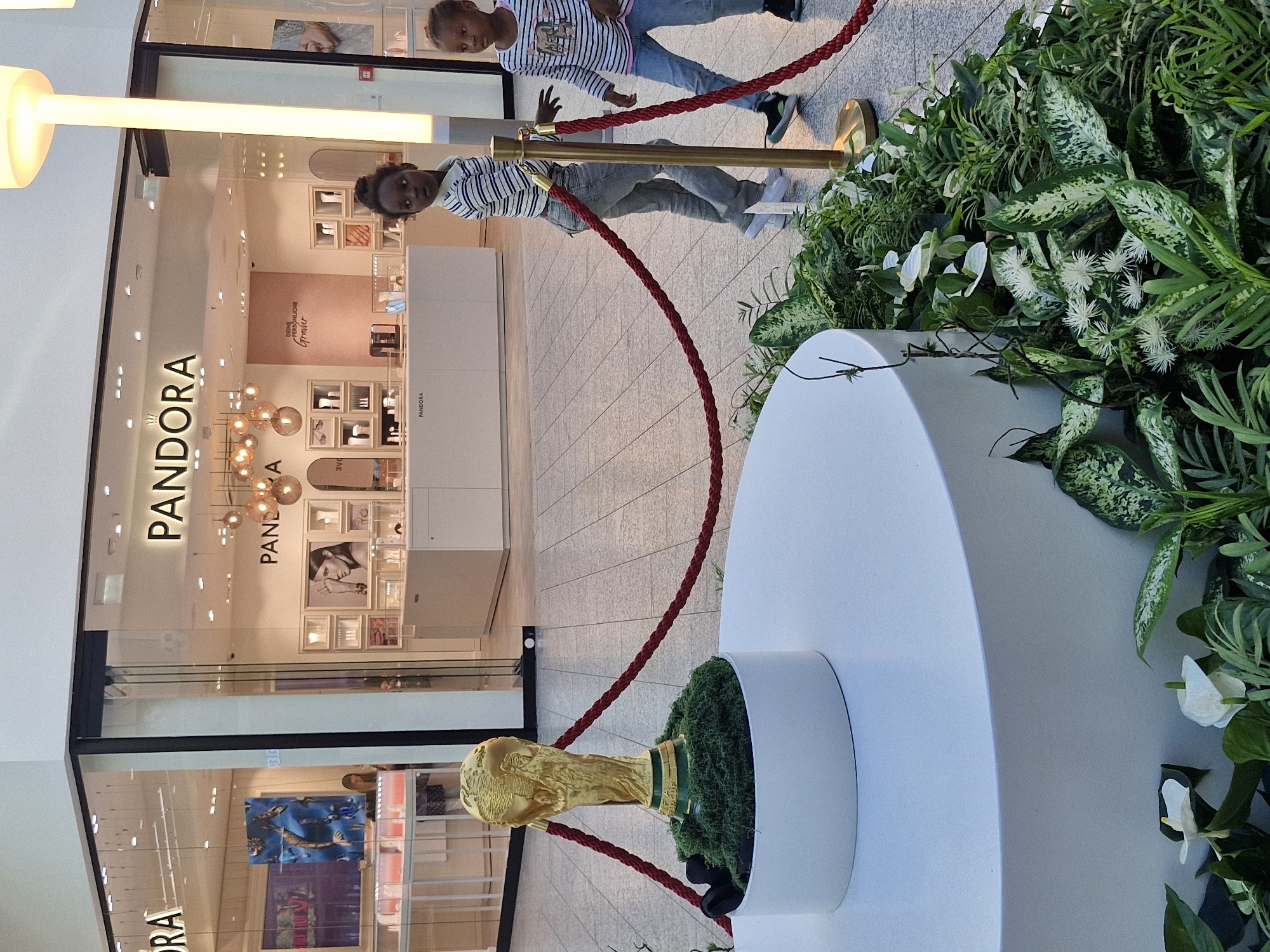 The image depicts the interior of a shopping mall in Bremen, Germany, featuring a Pandora jewelry store. In the foreground, a replica of the FIFA World Cup trophy is displayed on a white cylindrical pedestal, surrounded by lush green plants. A red rope barrier, supported by gold stanchions, delineates the display area. Two young girls, dressed in striped shirts and jeans, are visible near the barrier, one looking towards the camera and the other glancing to the side. The Pandora store's facade is visible in the background, with its prominent signage and display windows showcasing jewelry. The mall's floor is tiled, and ambient lighting illuminates the scene.