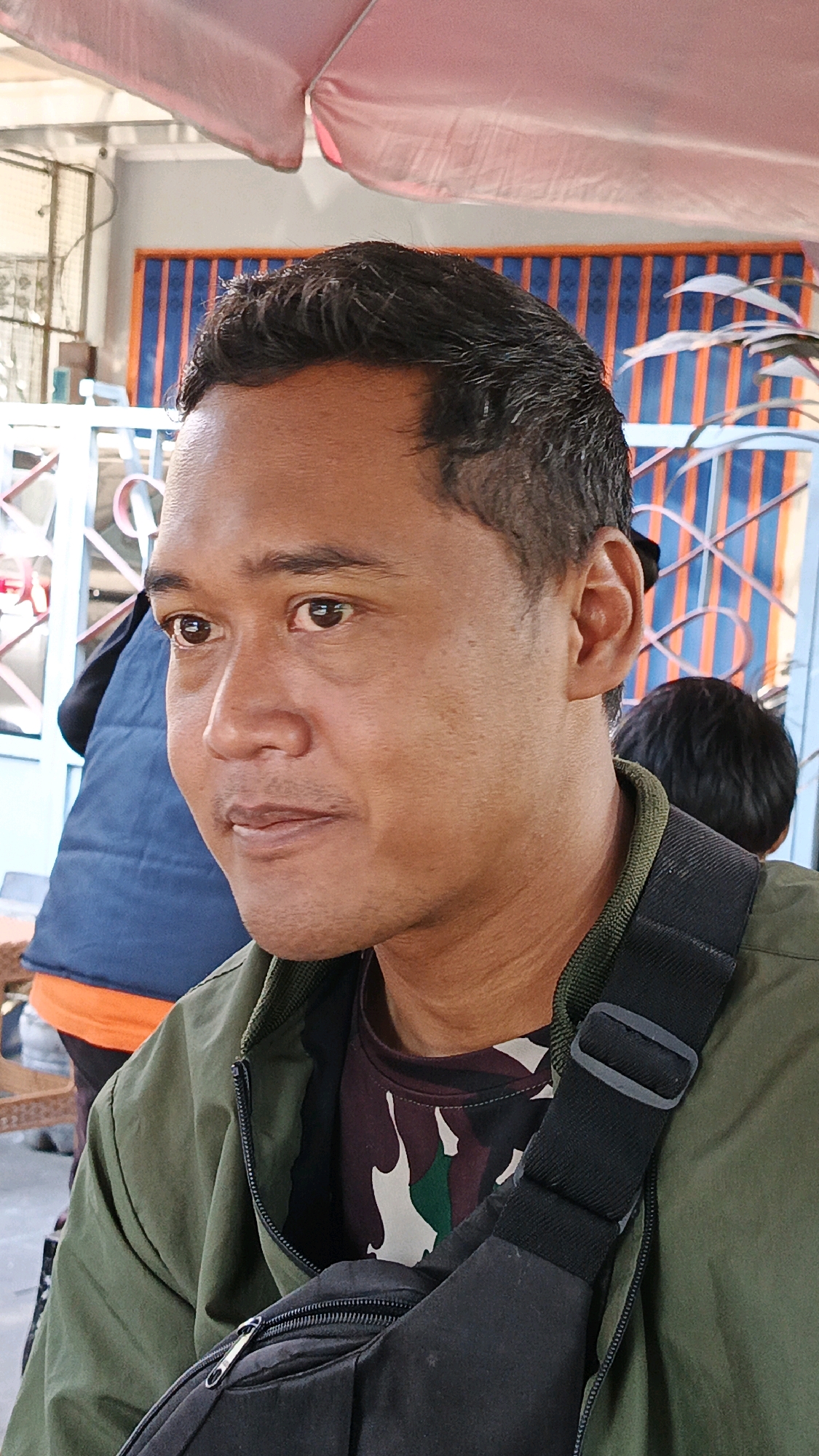 The image captures a close-up, profile view of a man outdoors, possibly in Surakarta, Indonesia, given the context. He has dark hair and medium skin tone, with a subtle mustache. He is wearing a green jacket over a camouflage-patterned shirt and a black strap that appears to be part of a bag or backpack is visible across his chest. His gaze is directed to the left of the frame, and his mouth is slightly open. In the background, a blurred figure of a person with dark hair is visible, along with a structure featuring vertical blue and orange stripes and a light-colored tent or canopy overhead. A metal fence or railing is also present in the background. The lighting suggests it is daytime.
