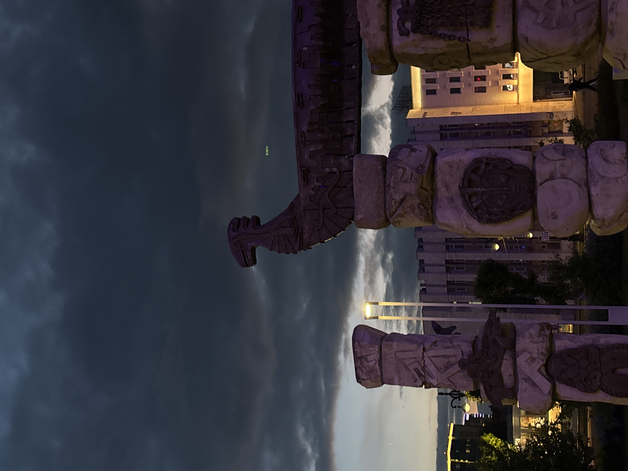 The image depicts the "Perm Gate" (Permskie Vorota) monument in Perm, Russia, at twilight or early evening. The upper two-thirds of the frame are dominated by a dark, heavily clouded sky, with a brighter, thinner band of clouds near the horizon on the left and a single bright point of light (likely a star or planet) in the upper left. A faint, diffuse green light pattern is visible in the upper right sky.
The monument itself, constructed from large, carved wooden logs, forms an archway in the lower half of the image. The logs are treated to appear dark and are illuminated with purple and blue artificial lighting. The horizontal top beam of the arch culminates on the left with a stylized head resembling a mythical beast, possibly a dragon or bird, featuring a prominent snout/beak. The supporting pillars below are composed of stacked, carved log sections. The central-right pillar displays a circular, mask-like face carving. The left pillar features various geometric patterns and a relief carving of a figure riding an animal.
In the background, multiple urban buildings are visible. On the far left, a modern multi-story building with illuminated windows rises above trees. In the center-right, a large building with a distinctive crenellated roofline and illuminated windows stands prominently. Further to the right, another building shows several illuminated windows, one with a distinct red light. Streetlights with bright, rectangular yellow-white lamps are present throughout the scene. Dark trees and bushes are interspersed around the base of the monument and foreground. A single person, silhouetted, walks on a paved surface towards the background buildings in the lower right quadrant. A white pickup truck is partially visible behind the walking figure.