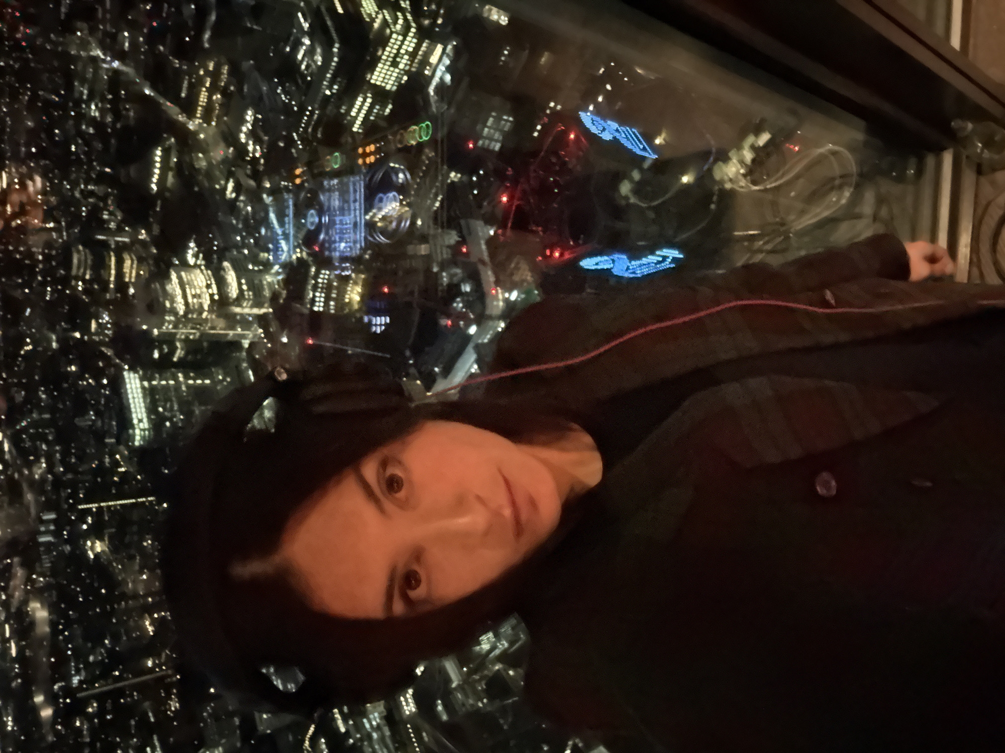 The photograph depicts a person with fair skin, dark hair, and brown eyes looking towards the camera. The person is wearing a dark shirt and a dark jacket or coat with a visible button. The background shows a nighttime cityscape with numerous lights, which appear to be viewed from a high vantage point, possibly through a window. Reflected in the window are two blue, spacecraft-shaped lights, possibly decorations, and other lights and structures. Based on the style of the architecture, street layout, and distinctive lighting, the scene seems to be taken from the Shard building in London, United Kingdom.