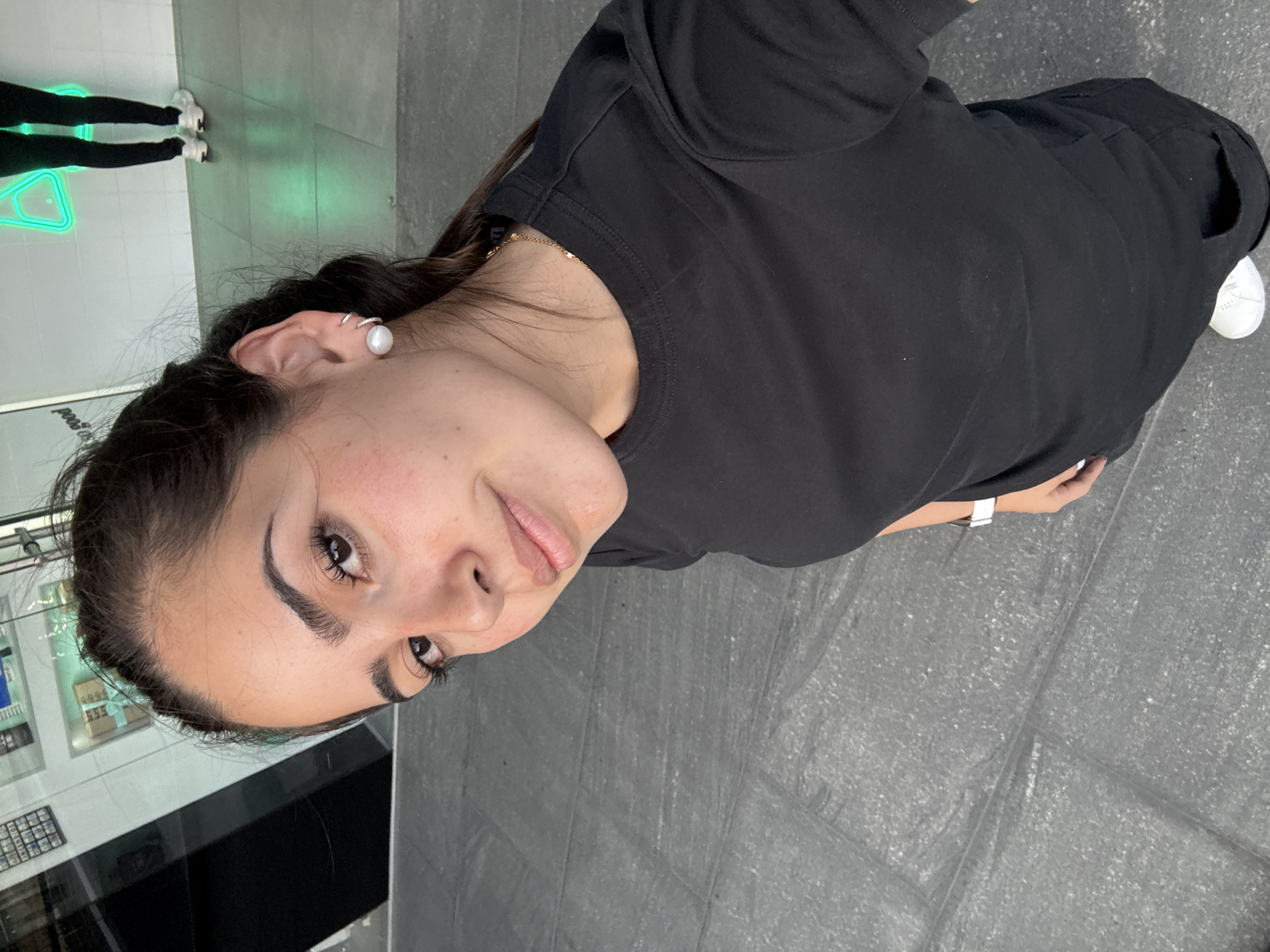 A young woman is standing indoors on a gray tiled floor. She is wearing a black t-shirt and black pants, and white sneakers are visible on her right foot. She is wearing a white wristwatch and a pearl earring is visible on her left ear. Another person is partially visible in the background with black leggings and white sneakers next to a neon light fixture on the glass. The woman is looking towards the camera. A display case is behind the woman on her left side. The location is San Bernardino Tlaxcalancingo, Mexico.