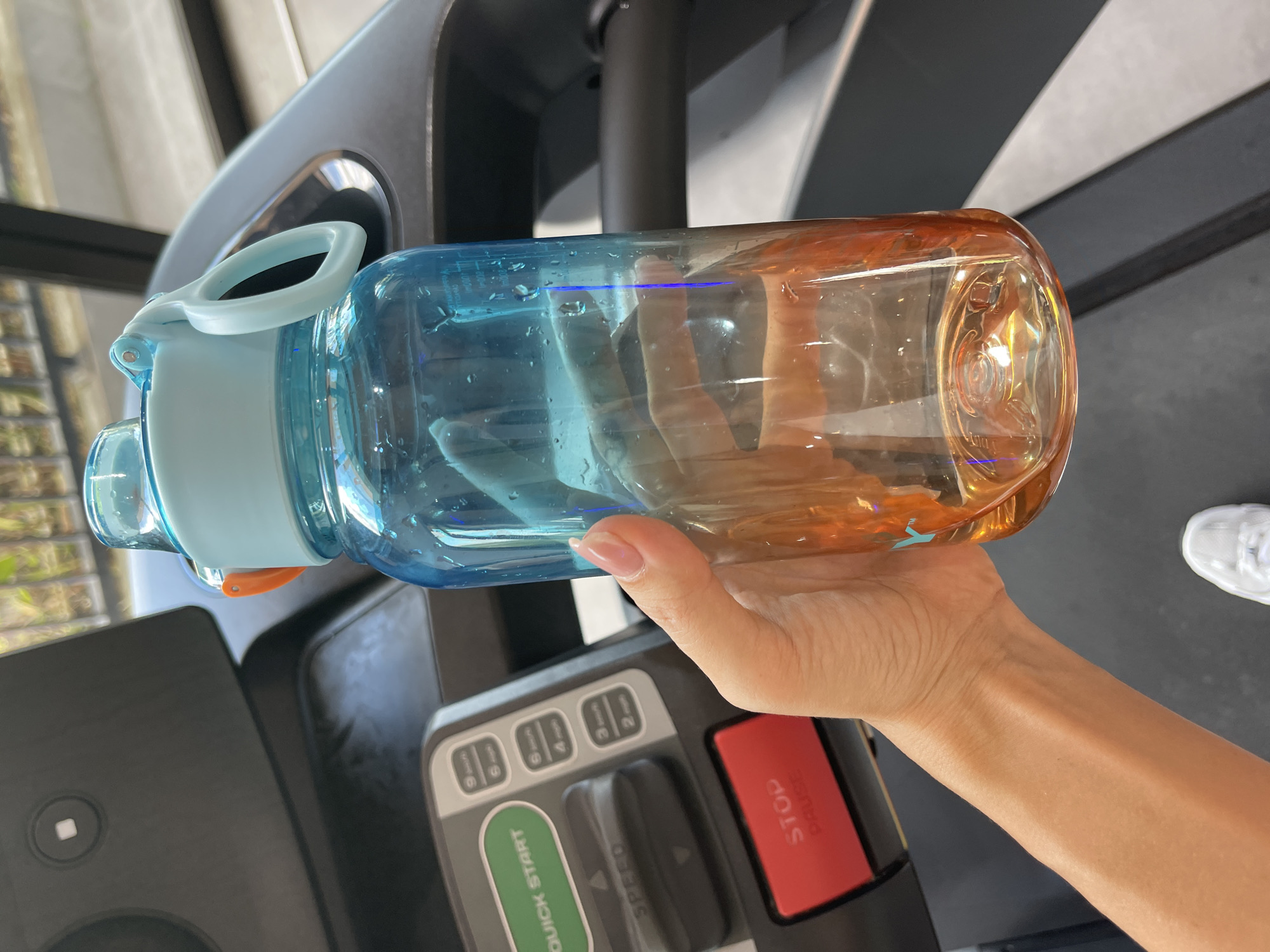An individual's hand is holding a blue and orange gradient water bottle with a light blue lid over a treadmill. The treadmill console is visible on the left side of the image, displaying buttons for "QUICK START", "SPEED", and "STOP/PAUSE". The water bottle has water droplets on the exterior. The background shows the upper part of the treadmill and a blurred outdoor environment. The scene is indoors.