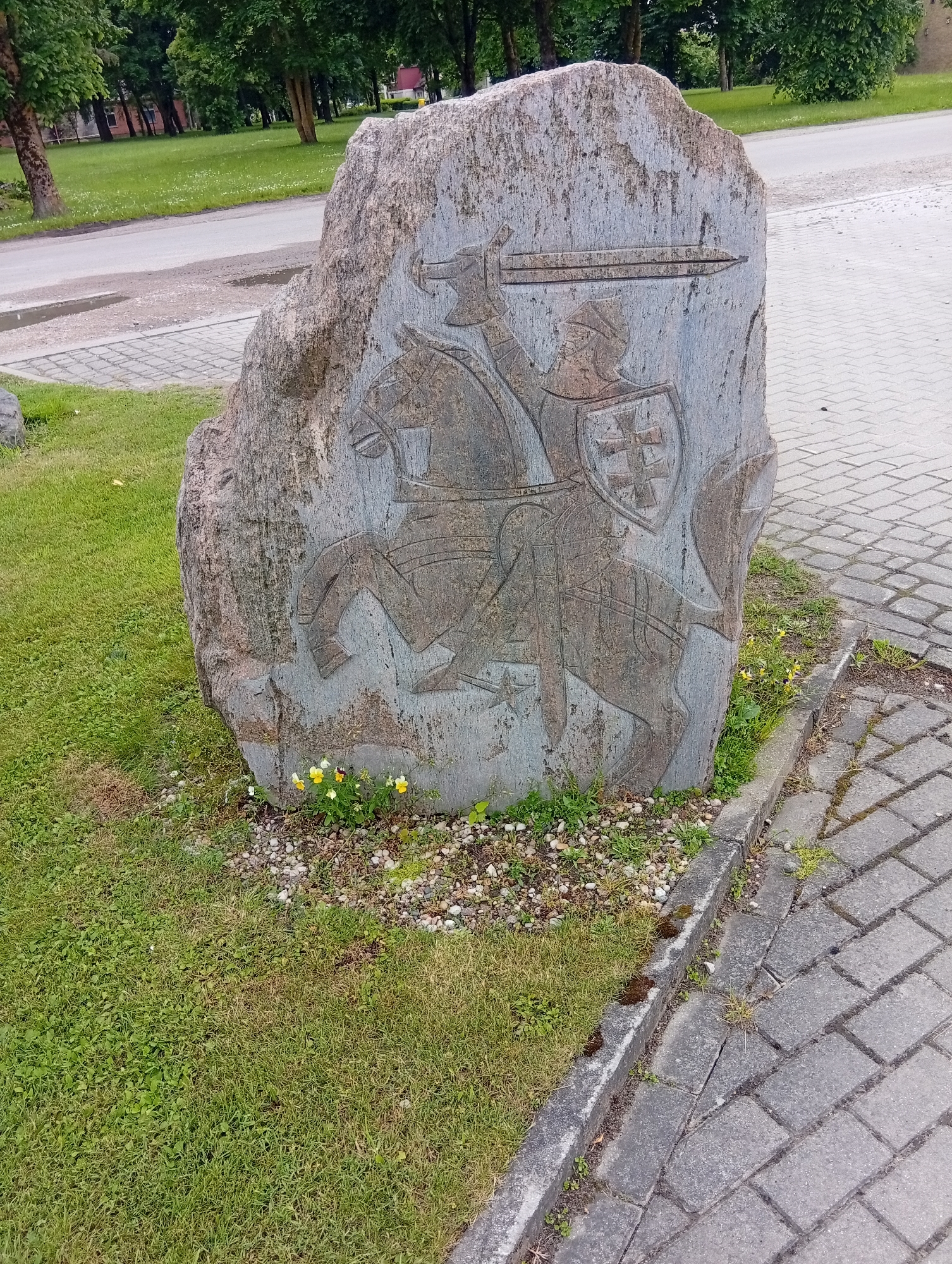A stone monument with a carved image of a knight on horseback is situated in front of a paved walkway and a grassy area with trees in the background. The knight is depicted wearing armor and holding a sword, with a shield bearing a cross symbol. Small yellow and white flowers are growing at the base of the monument. The monument appears to be located in the city of Micaičiai, Lithuania.