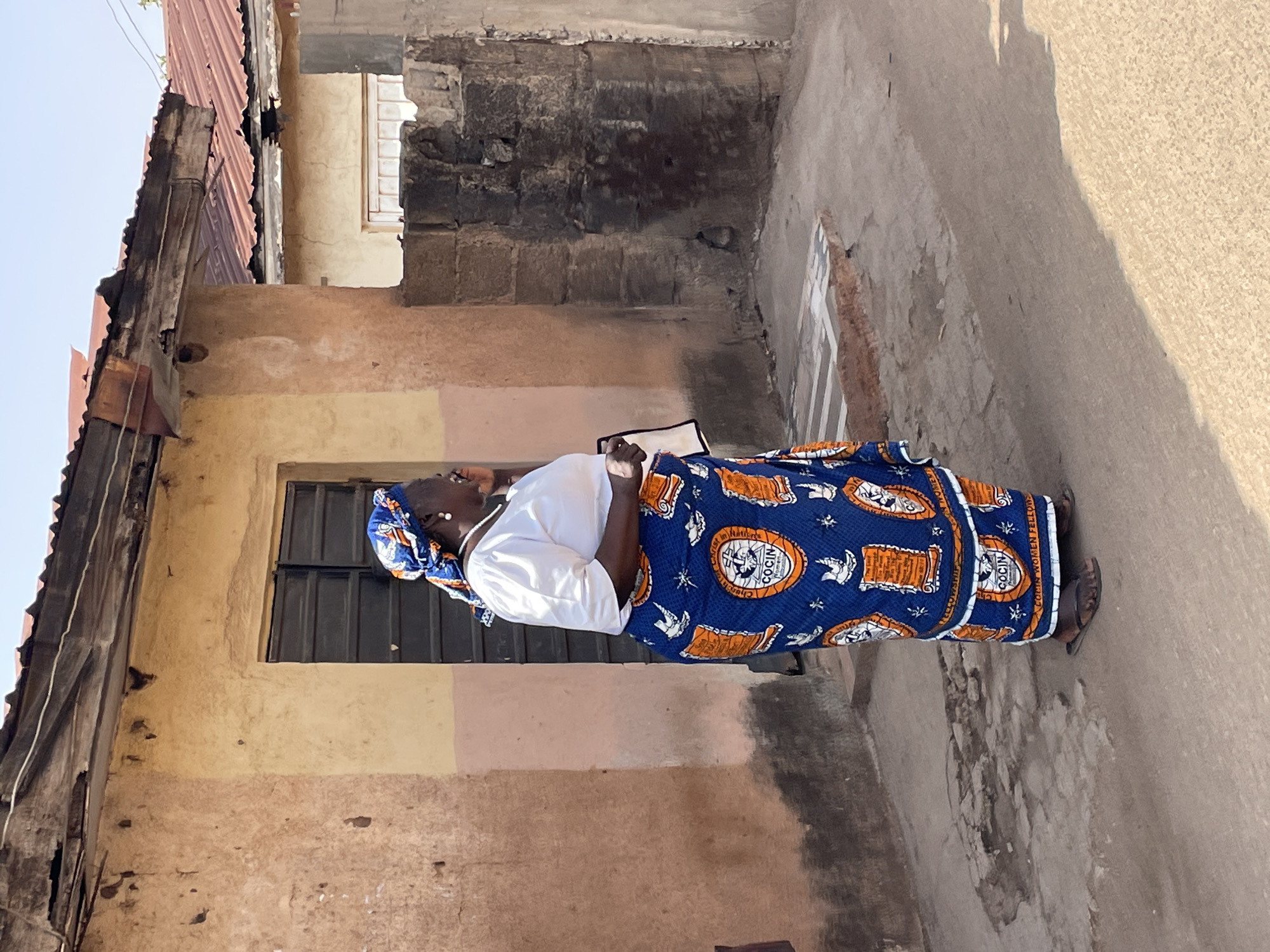 An outdoor, full-body shot captures an elderly woman standing on a narrow, unpaved path between two buildings under bright daylight. The image is oriented vertically, with the woman on the left side, facing towards the upper right. She wears a traditional blue and orange patterned skirt (wrapper) and a white short-sleeved top, with a matching blue headwrap. On her feet are simple sandals. She holds a small, light-colored object, possibly a book or tablet, in her hands.
The surroundings suggest a residential or community area. To her left, a building with textured, multi-toned walls (yellow, pink, brown) and a dark, shuttered window is visible. Above, a rusty, corrugated metal roof and exposed wooden beams are partially seen against a clear, light blue sky. To her right, a rough, earthy wall rises, casting a prominent shadow on the ground, indicating a sunny day. The ground itself is uneven and appears to be made of packed earth or concrete.
Notable details include the text on the woman's skirt. Circular motifs are visible, with one clearly stating "COCIN" at the bottom and "CHURCH OF CHRIST IN NIGERIA" around a central emblem, possibly a globe with religious symbols. Along the hem, the words "COCIN WOMEN FELLOWSHIP" are also visible. The architecture and traditional attire are consistent with the provided location context of Jos, Nigeria. The woman's expression is not clearly discernible, but her posture suggests she is calmly observing her surroundings or preparing for an activity.