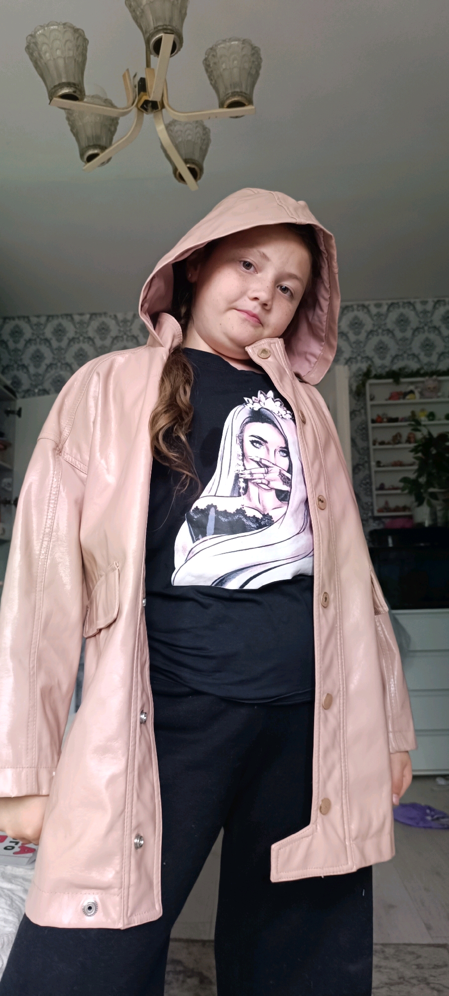 The image shows a young girl with light brown hair and freckles standing in what appears to be a bedroom. She is wearing a light pink hooded jacket over a black t-shirt with a graphic print of a woman's face wearing a crown. She is also wearing black pants. The background shows a patterned wallpaper, a white dresser with shelves filled with various items, and a light fixture on the ceiling. There are no discernible elements in the image that would suggest a location in the city of Chaikovsky, Russia. The scene appears to be an indoor setting.