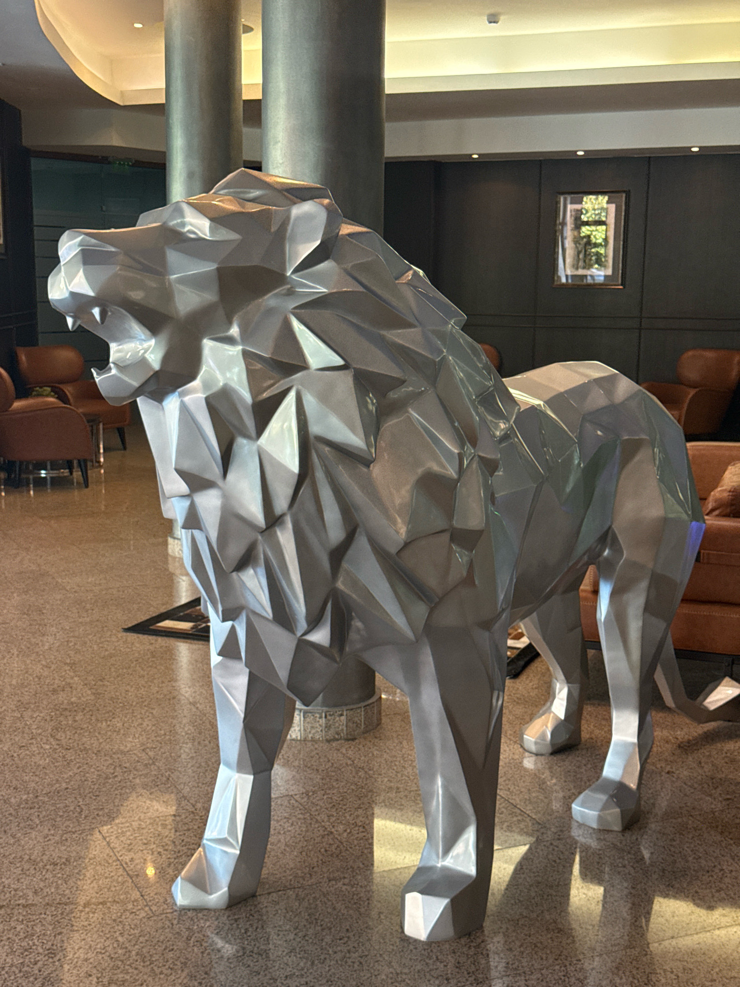 The image displays a metallic, polygonal sculpture of a lion in a hotel lobby. The lion is positioned centrally and faces left, with its mouth open. The sculpture has a reflective surface that catches the light, highlighting its geometric facets. In the background, there are large, cylindrical columns, a dark paneled wall with a framed picture, and upholstered chairs in a warm brown tone. The floor is a speckled terrazzo. The overall environment suggests a modern and sophisticated interior space. The location is identified as Sveti Vlas, Bulgaria.